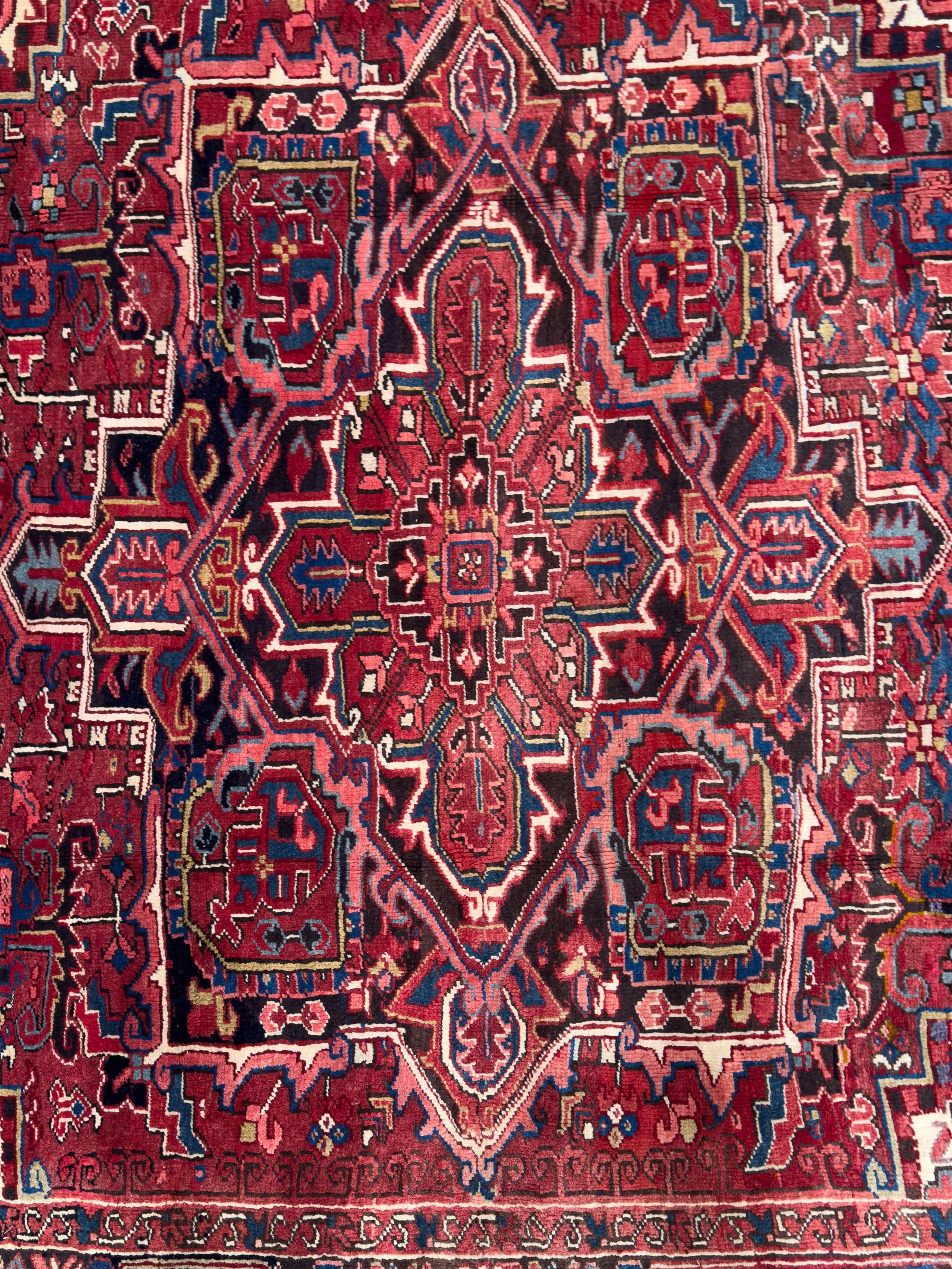 Northwestern Persian Heriz hand-knotted carpet, red madder field with a bold stepped medallion in indigo, ivory, and terracotta, flanked by angular floral motifs and stylized leaves, the spandrels set within a dark indigo border of rosettes and angular vine motifs between narrow guard stripes, 340cm x 245cm 