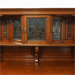 Early 20th century golden oak dresser sideboard, superstructure with three stained and leaded glass doors flanked by carved uprights above an open display shelf, the base with three drawers and geometric panelled cupboard doors, enclosing drawers with cutlery and bottle compartments, raised on short feet