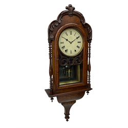 A 19thcentury American wall clock in a walnut case with crossbanding, with a carved pediment surround and arched glazed door flanked by two ring turned columns with pendants on an ogee plinth beneath, with a spring driven movement striking the hours on a bell, eight-inch painted dial with roman numerals, minute track and steel Maltese-Cross hands, with a spun brass bezel and winding collets, visible mock mercury pendulum with a reflective silvered mirror to the rear. With key and pendulum.   

