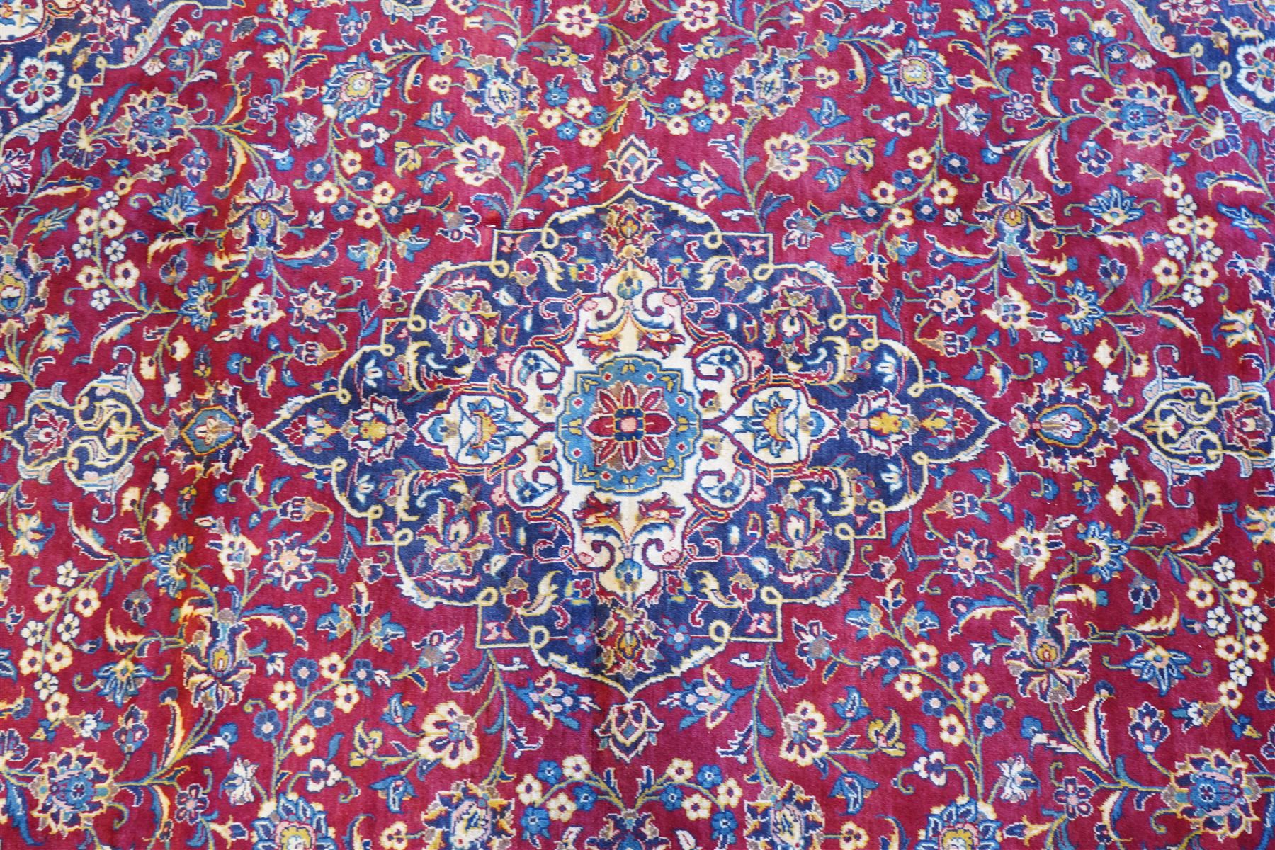 Central Persian Kashan hand-knotted carpet, crimson field with a central elaborate cusped medallion surrounded by elegant scrolling floral vine design incorporating stylized palmettes, rosettes and foliate sprays and similarly decorated spandrels enclosed by a dark indigo main border of floral arabesques between multiple guard stripes, cotton fringe ends 