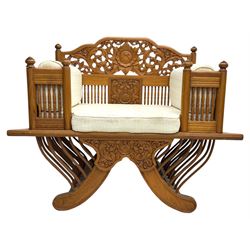 Early 20th century carved teak Howdah elephant chair, with pierced floral and foliate carved backrest, spindle gallery arm supports, cream upholstered cushions, splayed legs on a curved reeded base 