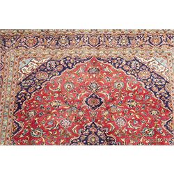 Persian Kashan hand-knotted carpet, madder red ground centred by shaped dark navy and ivory floral medallion with pendants, surrounded by scrolling vines, palmettes and flowerheads, dark navy spandrels, enclosed by dark navy floral border with multiple narrow floral guard stripes