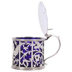 Early 20th century silver mustard pot and cover, of drum form the sides pierced with hound and birds in flight against leafy vines, the plain flat topped hinged cover with shell thumb piece opening to reveal a removable blue glass liner, hallmarked Joseph Rodgers & Sons, Sheffield 1929, including thumb piece H7cm