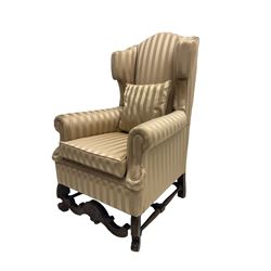 William and Mary style wingback armchair, upholstered in gold striped fabric, on walnut scroll carved supports joined by scroll and foliate carved middle rail, turned H-shaped stretchers, on castors