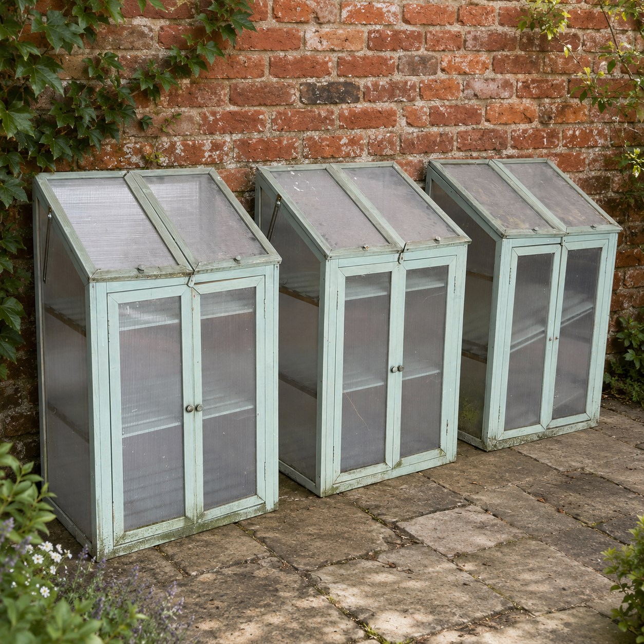 Three green-painted timber cold-frame greenhouses or growhouses, each with twin hinged sloping polycarbonate lids above a pair of panelled doors enclosing slatted shelves