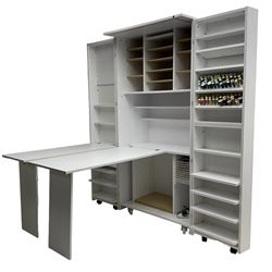 Large white finish sewing cabinet, enclosed by two doors fitted with thread holders and shelves, the interior fitted with shelves and sliding trays, with fold down work surfaces 