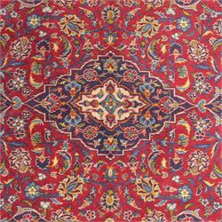 Central Persian Kashan hand-knotted rug, crimson field, profusely decorated with scrolling vines and palmettes, floral sprays around an ivory and indigo lobbed medallion enclosed by spandrels within an indigo border of arabesques between multiple guard stripes  