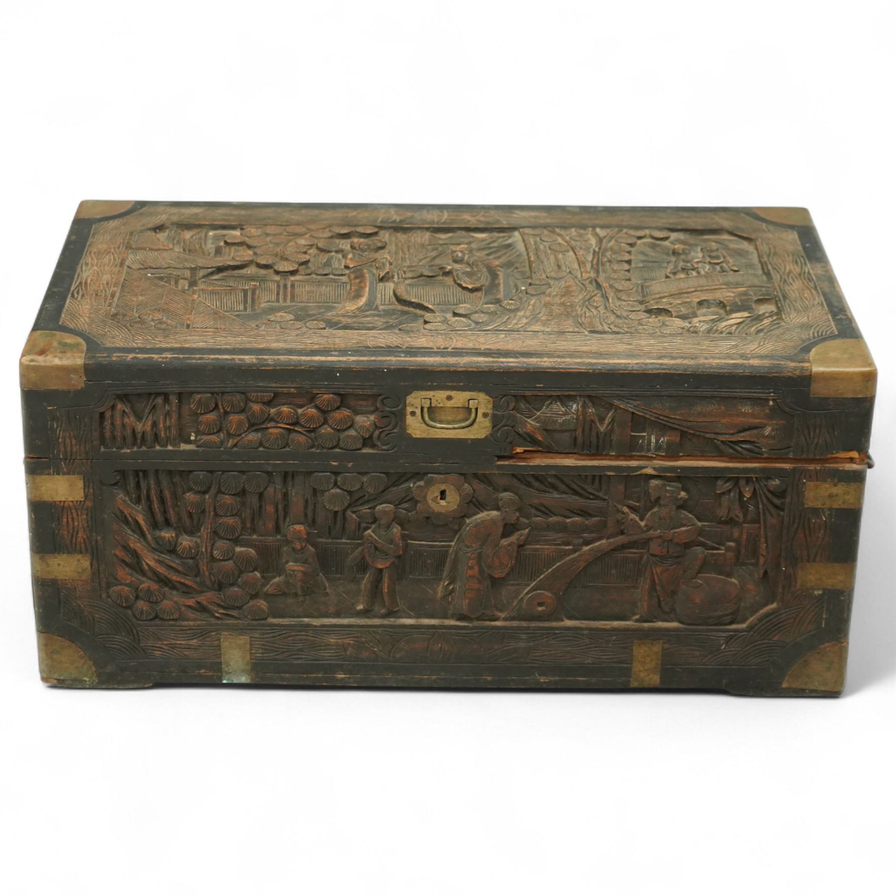 Early 20th century Chinese brass bound camphor wood coffer, rectangular form, the hinged lid and sides deeply relief carved with figures in landscape scenes within geometric borders, fitted with brass corner mounts and recessed carry handles
