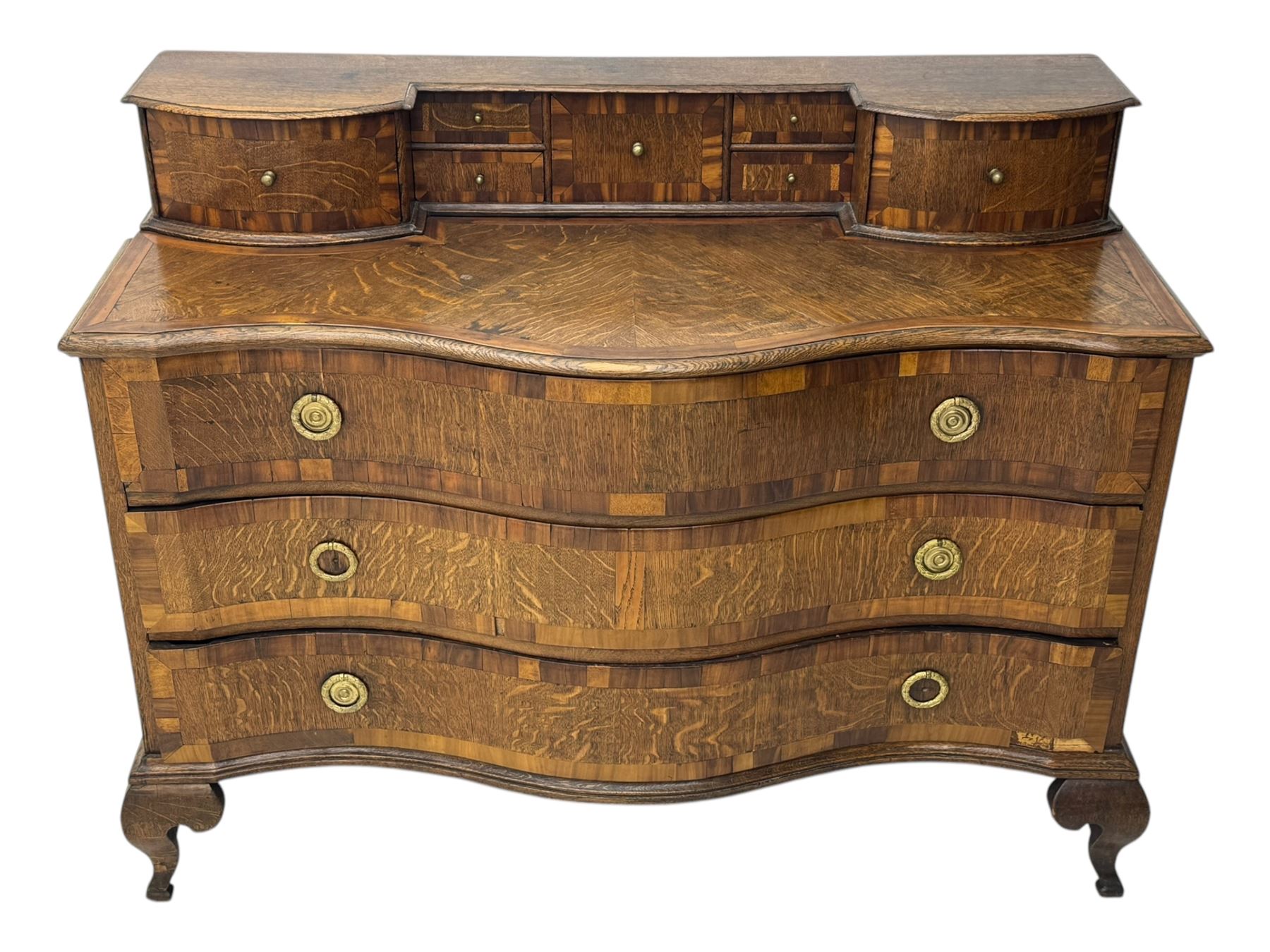 Late 19th century continental oak commode, serpentine fronted with parquetry and crossbanded decoration, the raised superstructure back fitted with a central bank of five recessed drawers flanked by concave drawers, above three full-width drawers with brass ring handles, raised on cabriole supports
