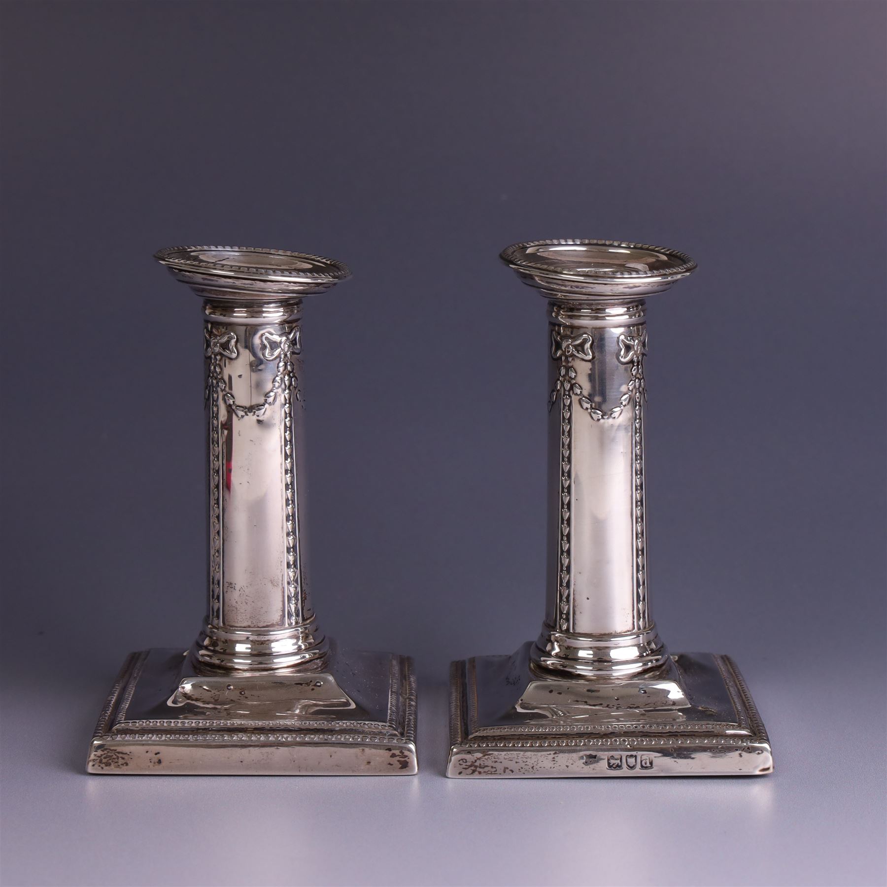 Pair of late Victorian silver mounted candlesticks, the stems embossed with classical ribbon and swag design, upon weighted stepped square bases, hallmarked London 1899, maker's mark worn and indistinct, H12cm