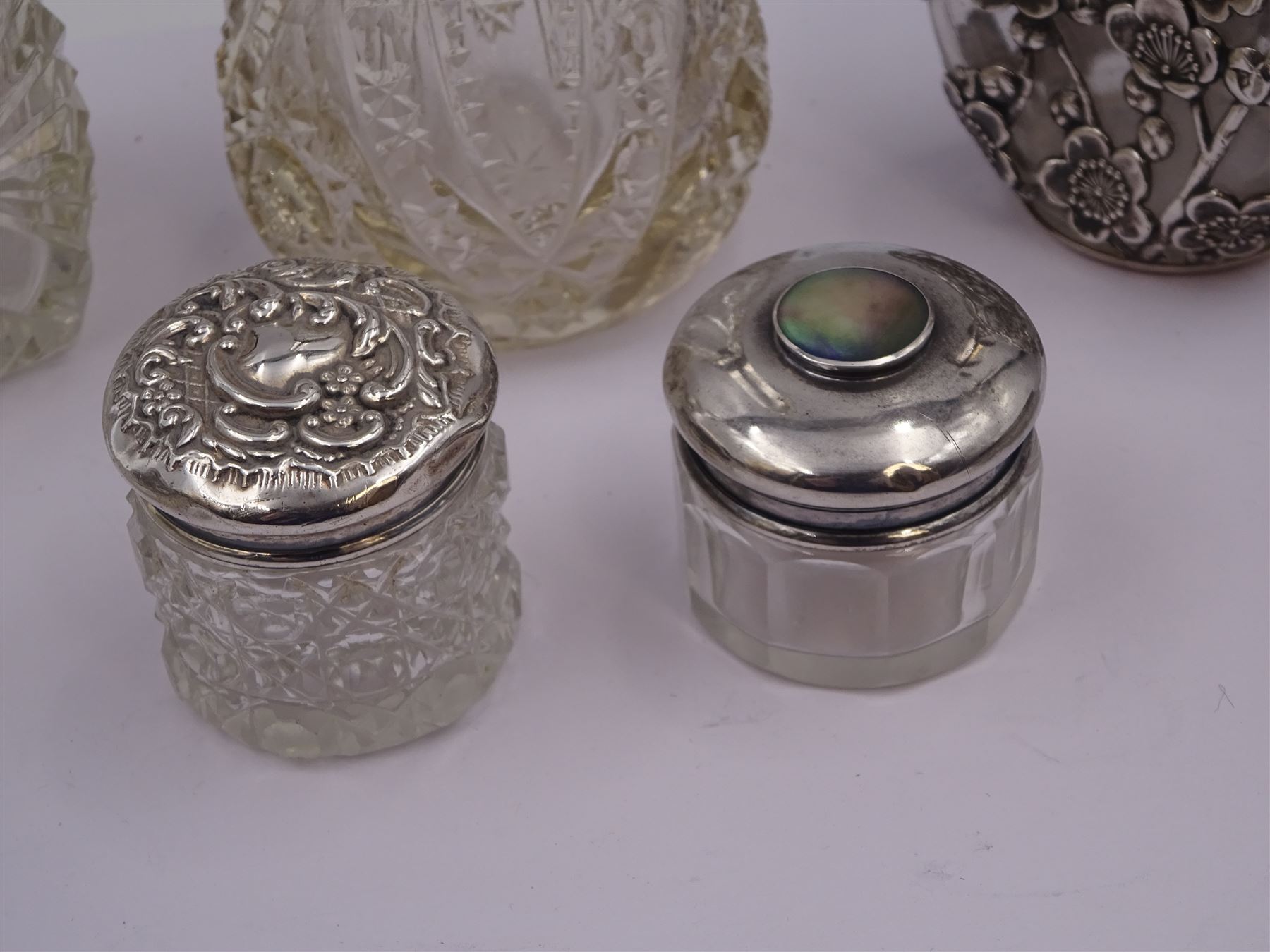 Two silver mounted dressing table jars, one in the form of a heart, both with embossed silver covers and cut glass bodies, hallmarked, together with two smaller examples, one set with mother of pearl, three silver mounted glass scent bottles, including an example with prunus blossom decoration, stamped Sterling, tallest bottle H17cm
