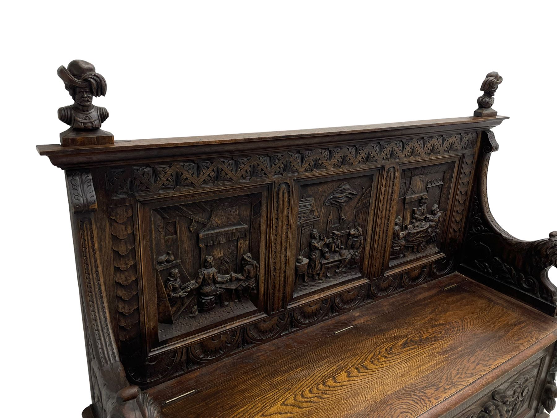 19th century heavily carved oak settle, the moulded upper rail over triangular foliage carved frieze, three Flemish style carved panels depicting tavern scenes, acanthus carved uprights terminating to lion carved arms, hinged box seat, the panelled front carved with lion masks and extending scrolled acanthus leaves, on moulded skirt base