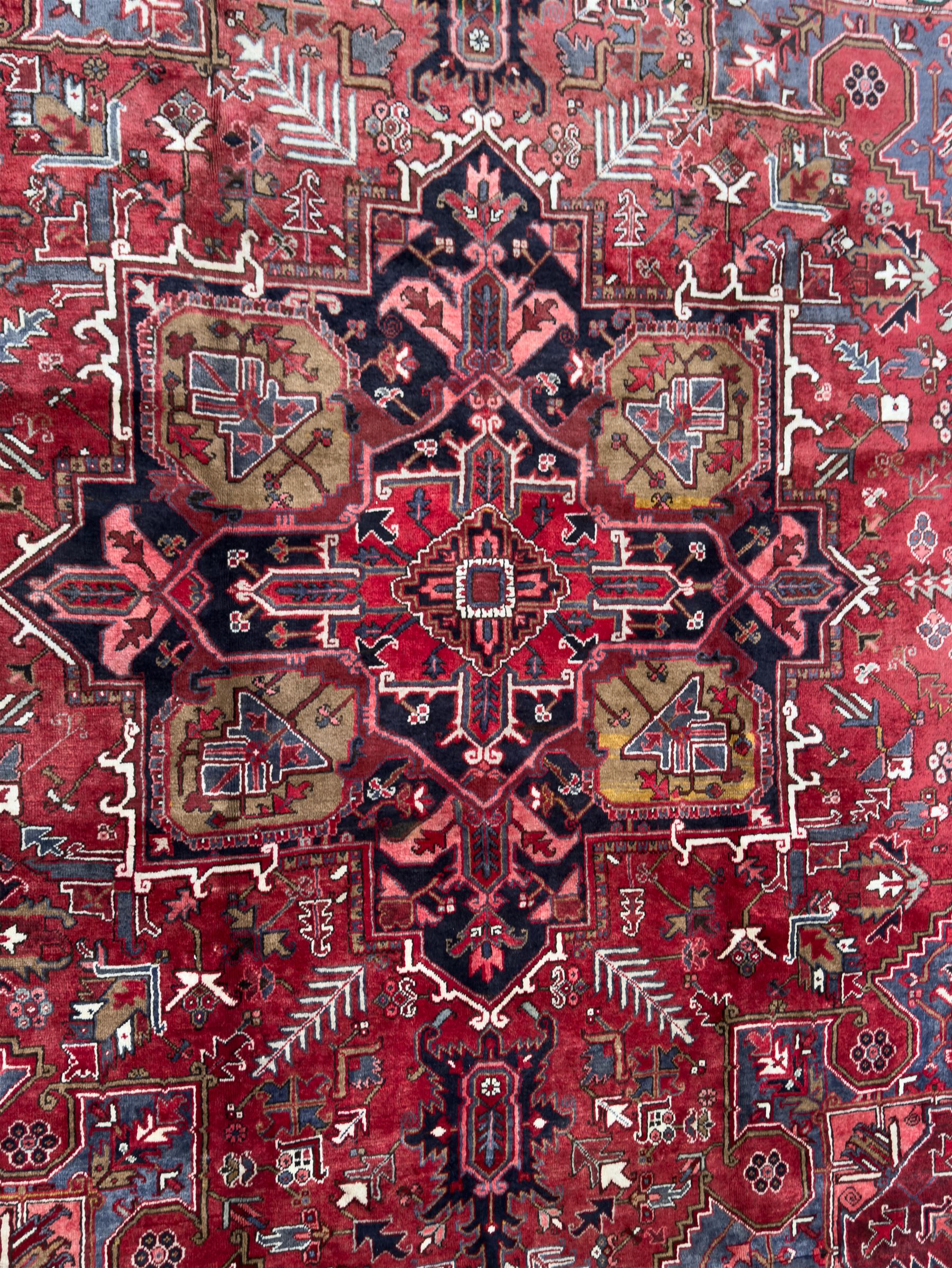 Northwestern Persian Heriz hand-knotted carpet, madder red field with a bold stepped medallion in indigo, ivory, and ivory, surrounded by angular floral motifs and stylized leaves, and the spandrels set within a dark border of rosettes and angular vine motifs, 335cmx252cm