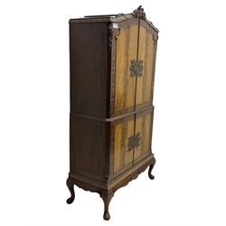 Early 20th century walnut veneered cocktail cabinet, shaped and carved foliate pediment above a pair of arched panelled doors with figured veneers and ornate cast escutcheons, interior is mirrored on the base and back, slide out mirrored shelf, flanked by foliate carved uprights, over a conforming lower cabinet, all raised on shaped apron and scroll carved cabriole feet