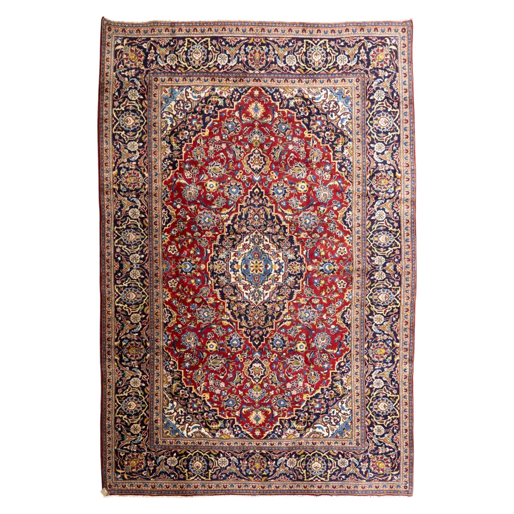 Central Persian Kashan hand-knotted rug, rich red field with a central lobbed medallion surrounded by finely drawn scrolling floral sprays, palmettes, and rosettes, enclosed by a broad dark blue floral border with multiple guard stripes and cotton fringe ends 