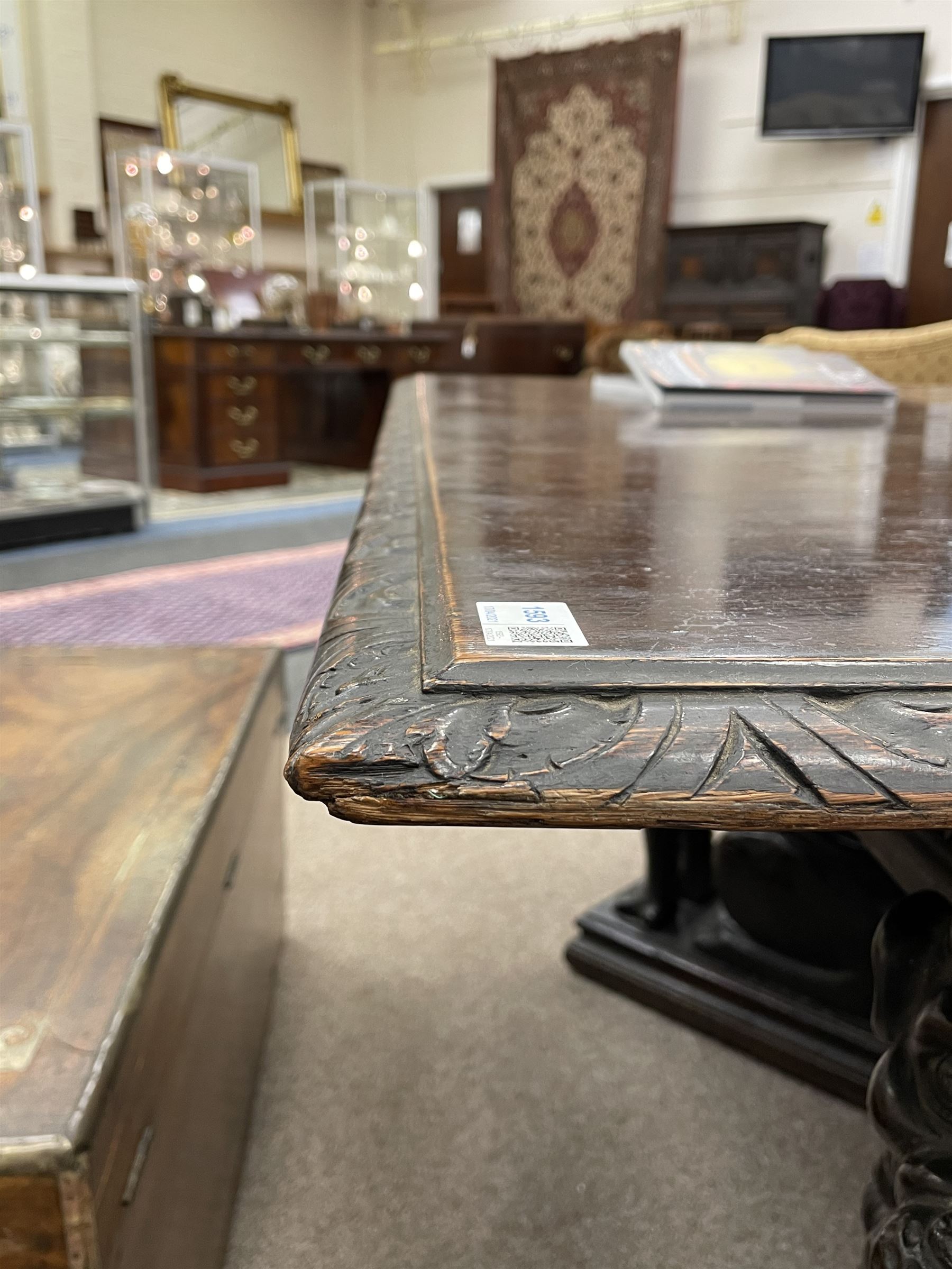 Large 19th century carved oak library table, the rectangular top with lunette carved edge over cartouche panelled and carved  scroll frieze, the central tapering rectangular support relief carved with masks and garlands, on four carved seated lion supports at each corner, on flat angular stretcher with recessed castors stamped ‘Bartholomew & Fletcher, London’