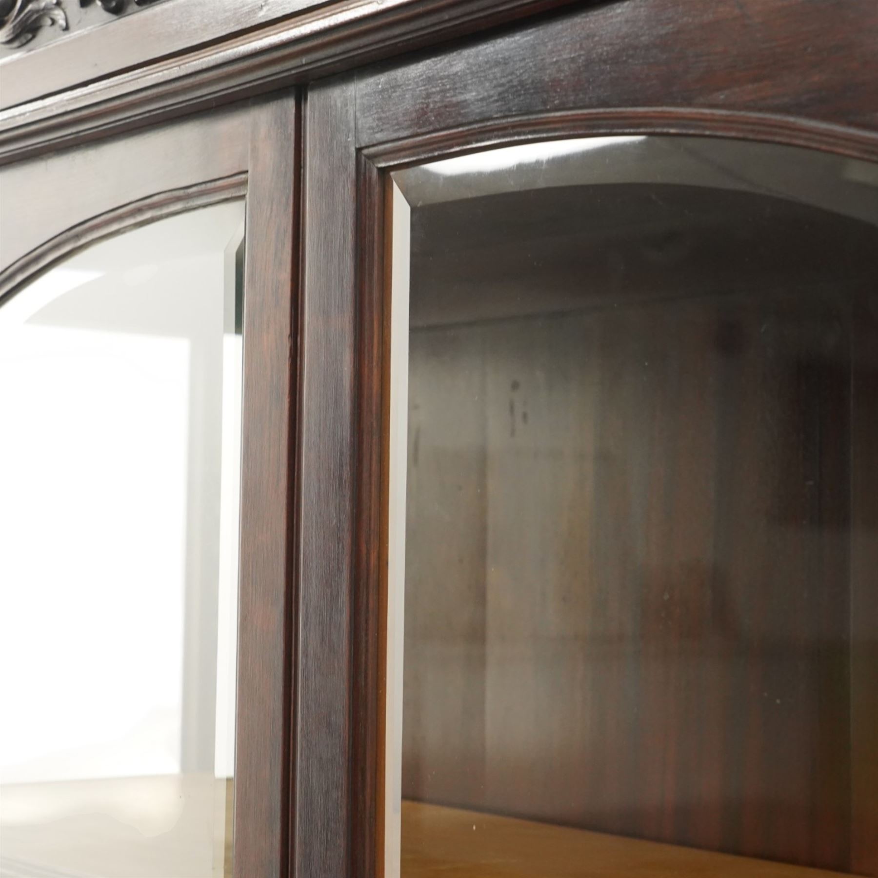 Large and impressive 19th century heavily carved mahogany display cabinet, the ornate pediment carved with a central scalloped and foliate rocaille motif with extending acanthus leaves and egg-and-dart moulding flanked by acanthi finials, over a central cartouche with carved bow-and-arrow motif and trailing scrollwork, the bookcase enclosed by two glazed doors with bevelled glass sides, the base with a gadrooned upper edge over a central rocaille ornament with scrolling acanthus leaves, the panelled frieze with further foliate motifs, raised on large carved snarling lion mask supports with floral garlands and claw feet, united by a concave rectangular platform base with gadrooning egg-and-dart moulding