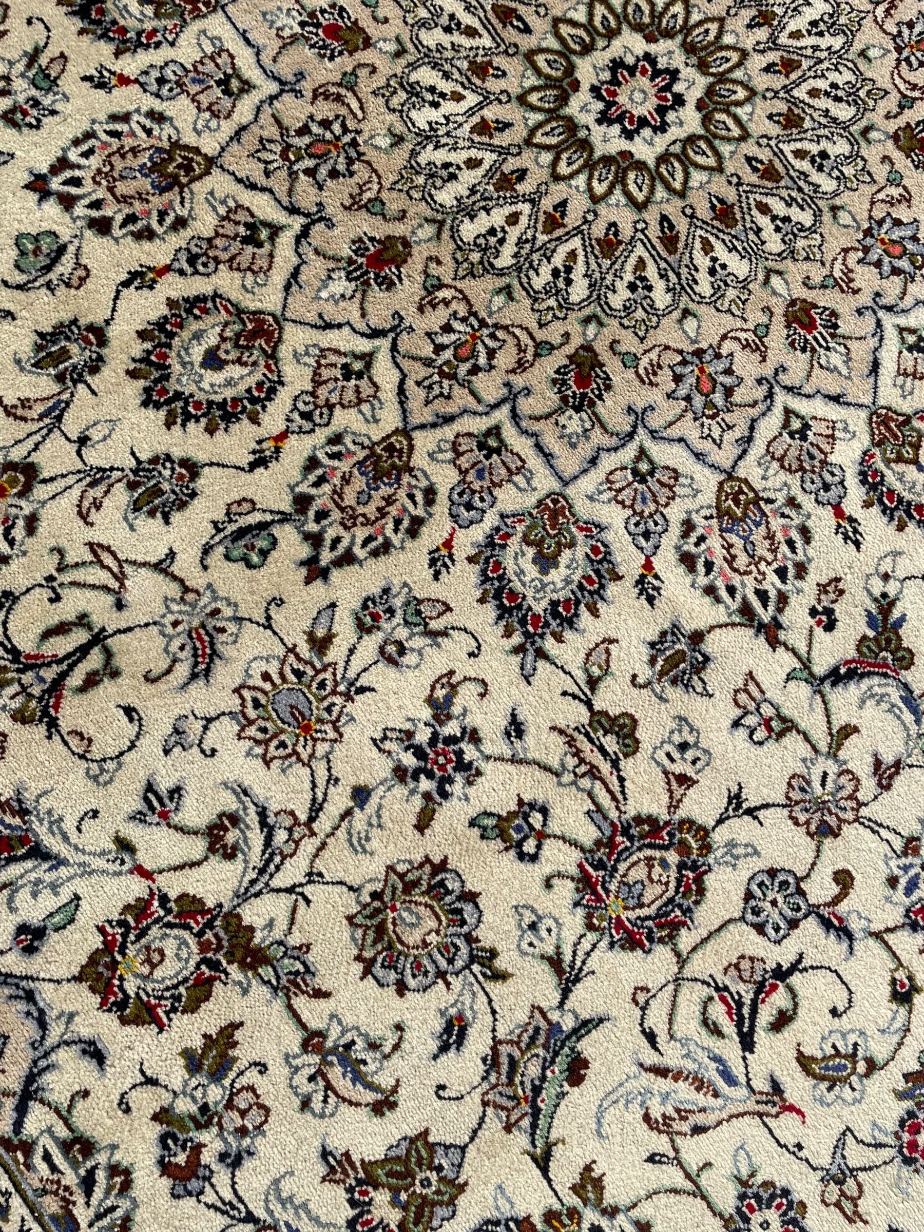 Persian Kashan ivory ground carpet, central rosette medallion with projecting palmettes, densely decorated field depicting trailing leafy branches and stylised plant motifs, the border with similar scrolling pattern, within floral pattern guard stripes 