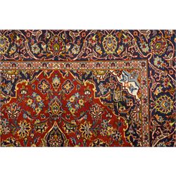 Central Persian Kashan hand-knotted rug, deep red field with central cusped ivory medallion with an all-over arrangement of scrolling floral vines, palmettes and flowering sprays within conforming spandrels, principal navy border with a continuous meandering vine of palmettes and stylized floral motifs, enclosed by multiple narrow guard borders, with cotton fringe ends 