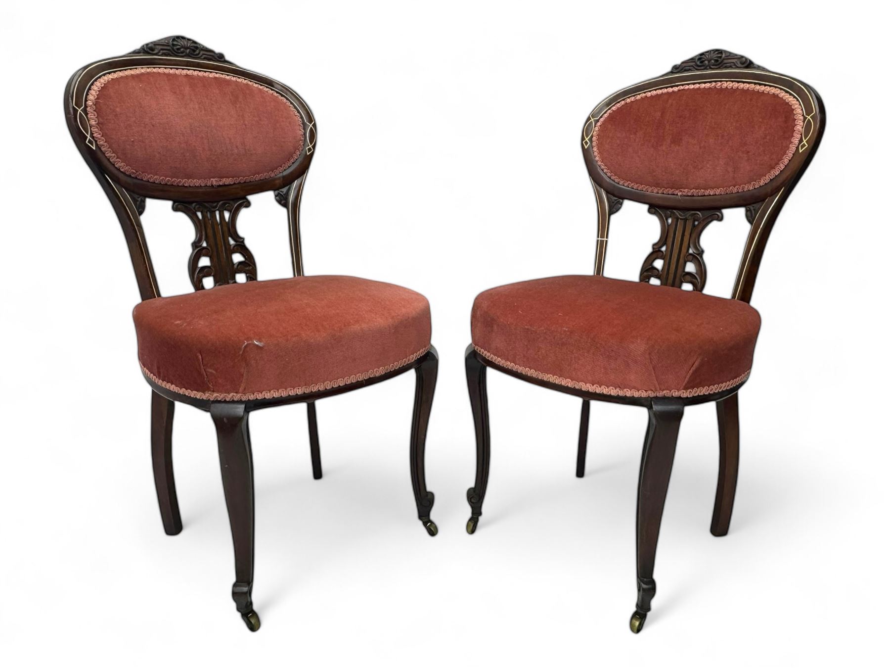 Pair of Edwardian inlaid rosewood salon chairs, upholstered in dusky pink fabric with gimp trim, oval backrest with carved crest rail with central shell motif and pierced splat, on cabriole supports terminating in castors