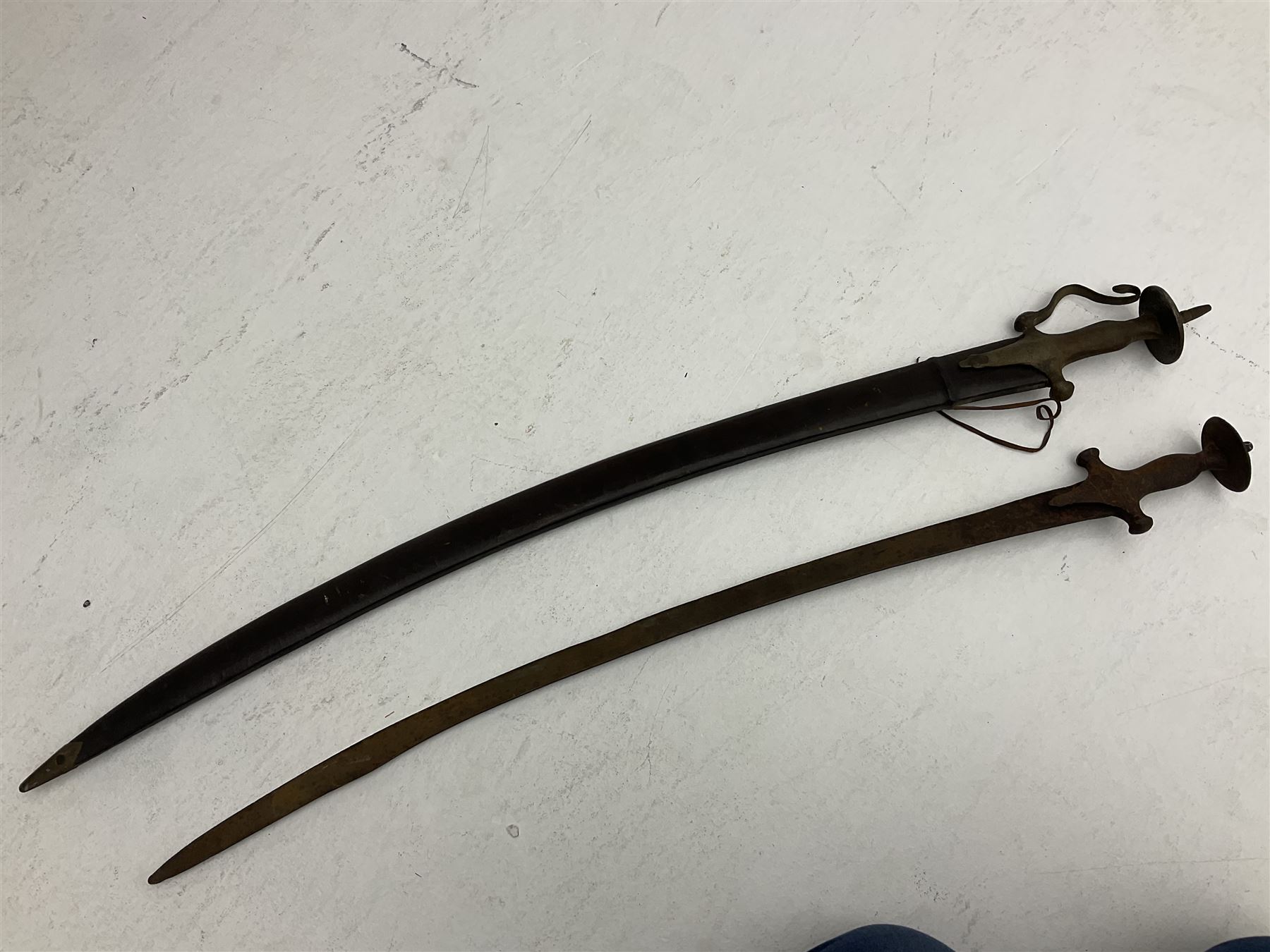 Late 19th/early 20th century Indian Tulwar sword with 81cm plain curving steel blade and iron hilt with extended langets, knucklebow and spiked pommel; in leather covered scabbard L100cm overall; and another Tulwar lacking scabbard in almost relic condition (2)