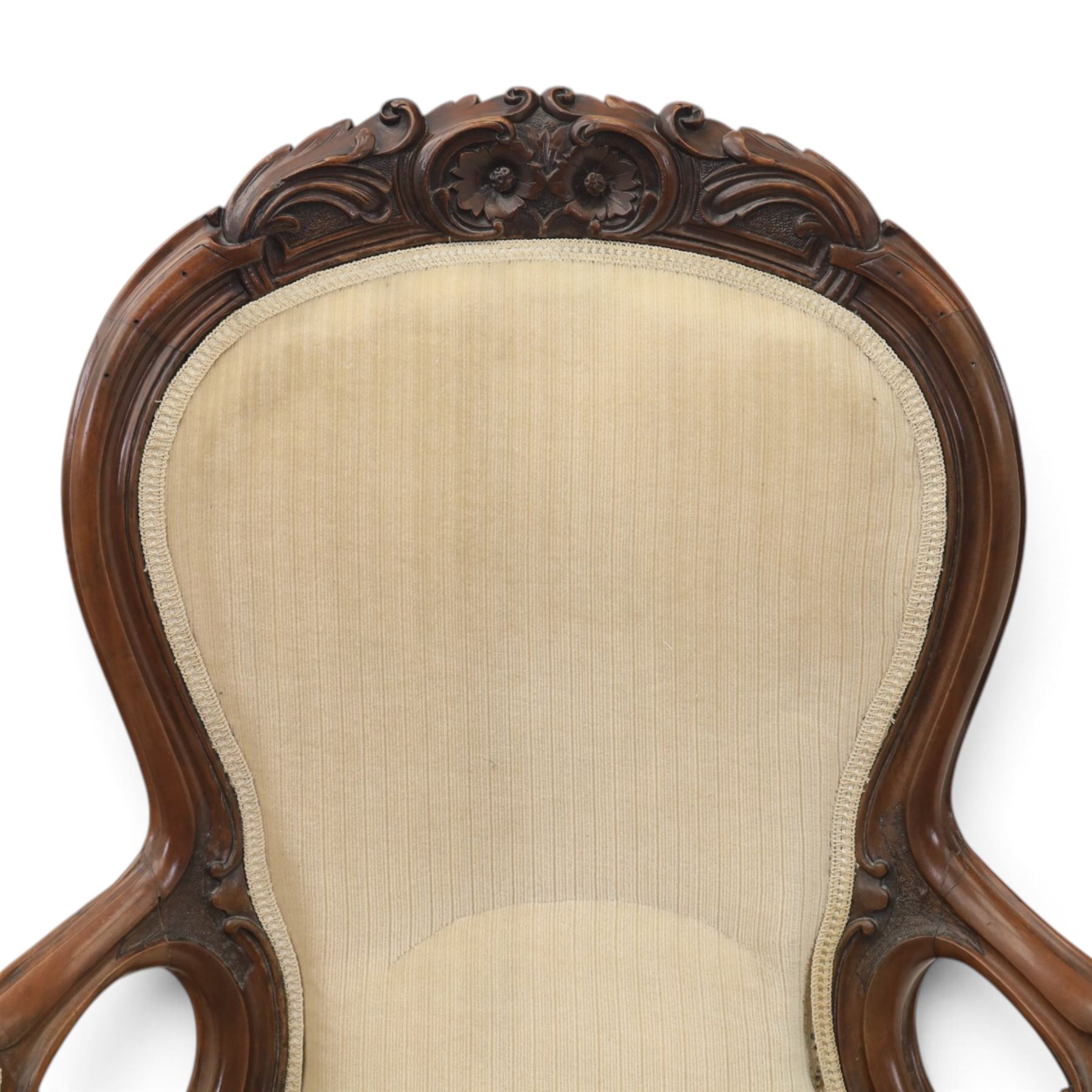 Late Victorian walnut open armchair, the carved shaped cresting rail centred by scrolls and foliage above a padded serpentine back and overstuffed seat upholstered in cream striped fabric, with padded scroll arms on open carved supports, raised on cabriole supports with carved knees and ceramic castors
