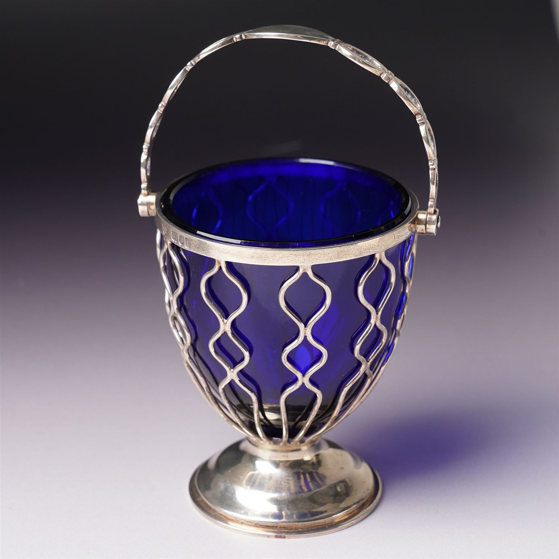 Edwardian pierced silver sugar basket with swing handle and blue glass liner H9cm London 1906 Maker Haseler Bros. and another decorated with floral swags Chester 1912 Maker  (2)