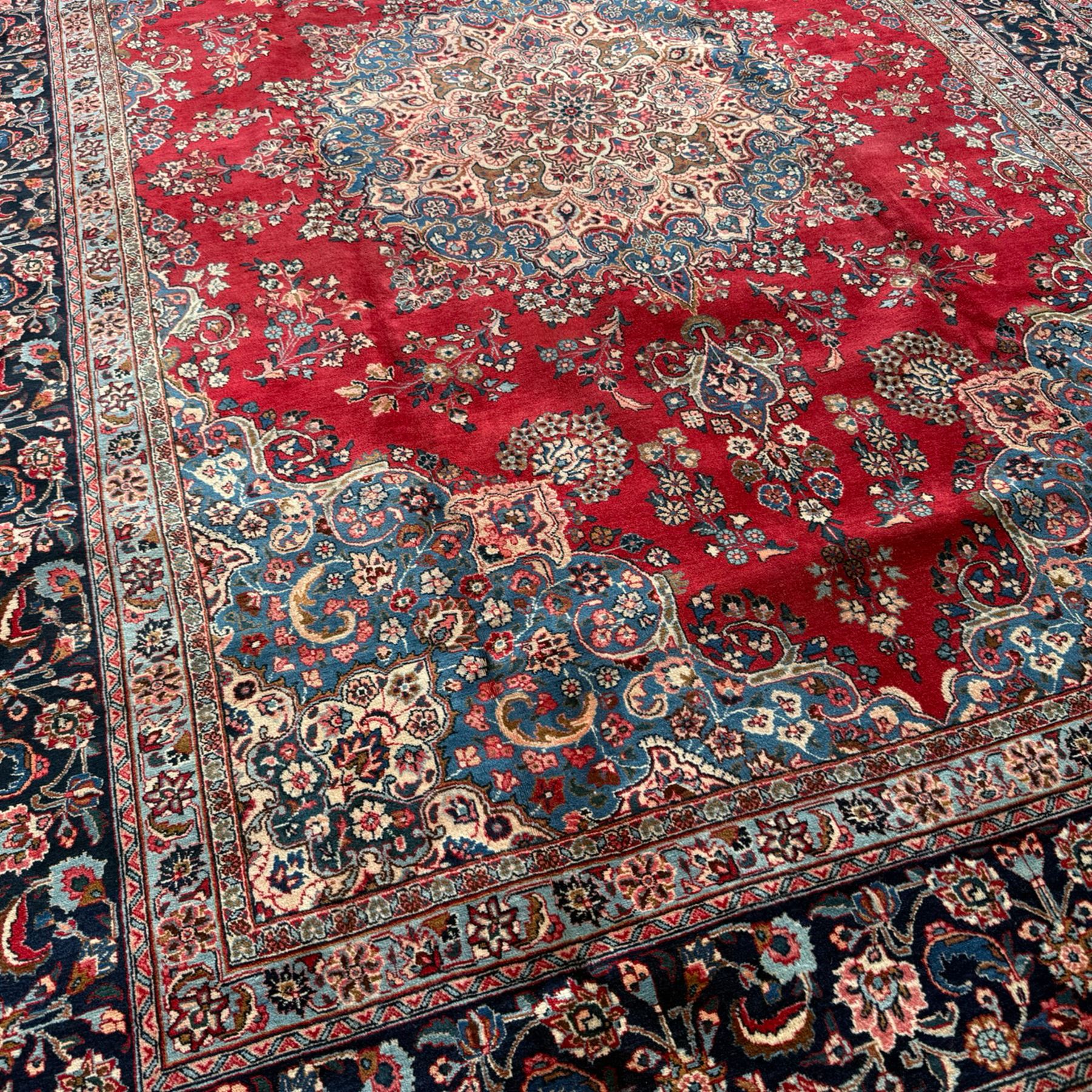 Meshed Persian rug, red, indigo, and teal background with repeating floral vine around a symmetrical floral medallion with bold palmettes, central ivory radiating floral centrepiece, and spandrels, indigo and teal border of delicate scrolling palmette and leafy floral vines