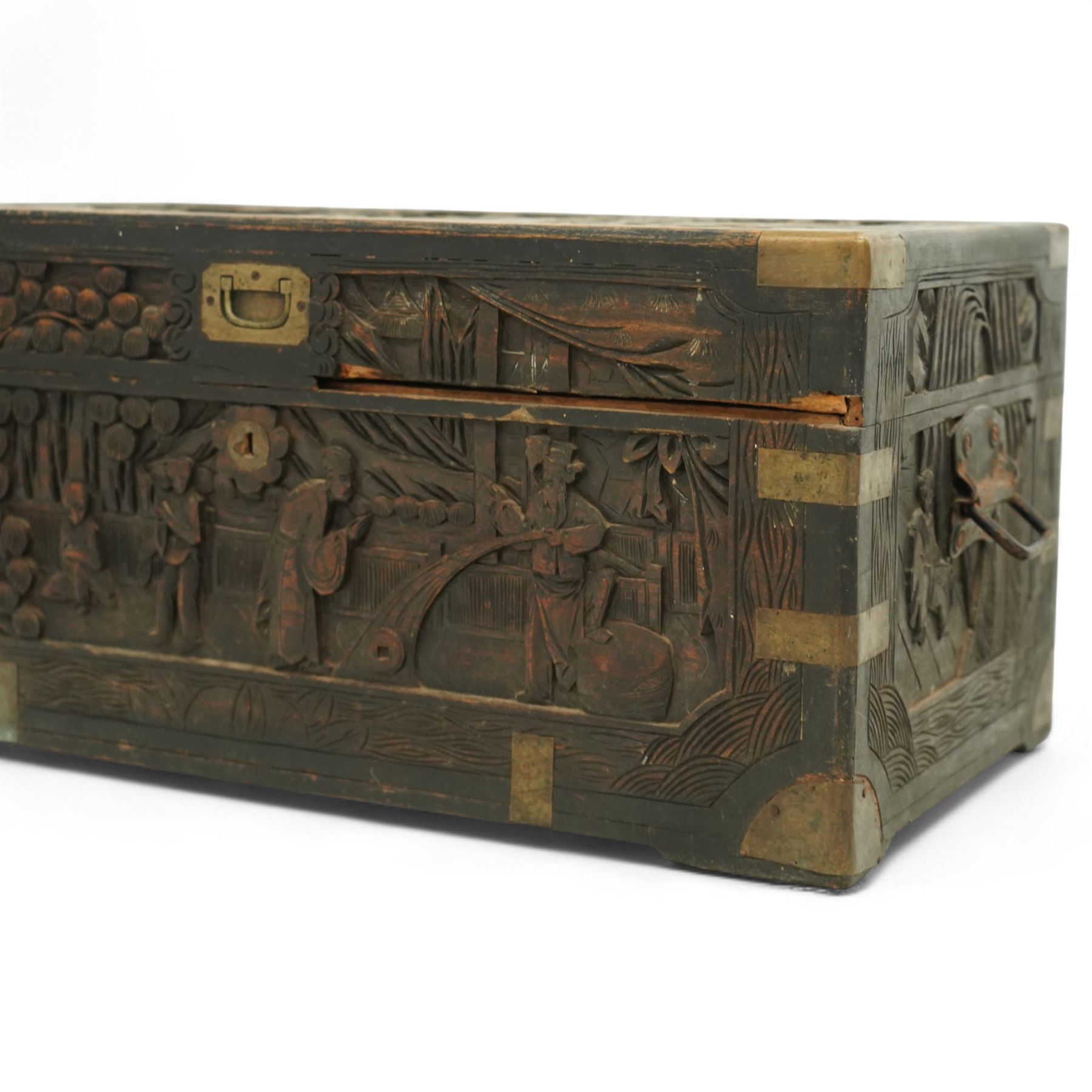Early 20th century Chinese brass bound camphor wood coffer, rectangular form, the hinged lid and sides deeply relief carved with figures in landscape scenes within geometric borders, fitted with brass corner mounts and recessed carry handles