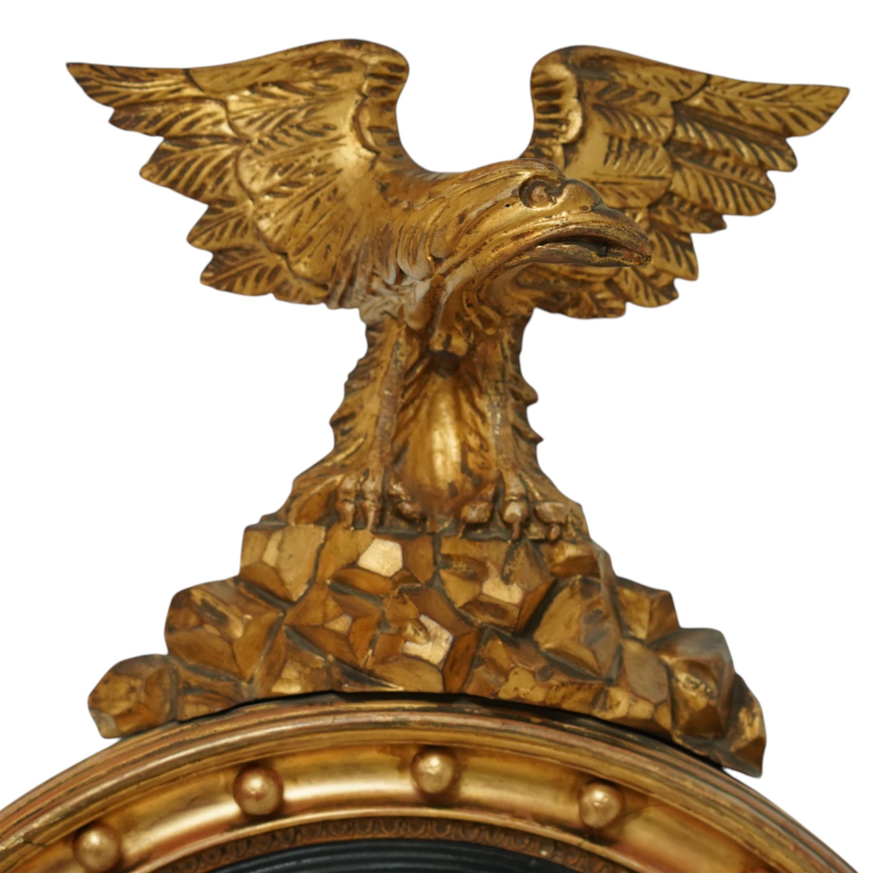 Regency period eagle mounted giltwood convex mirror, surmounted by a finely carved giltwood eagle standing on rocks above the circular mirror with the original mirror plate and surrounded by a fluted ebonized slip and the convex frame with gilt ball decorations, the back with label for ‘T.G. Woof Ltd, manufacturing cabinet makers, Devonshire Street, Sheffield