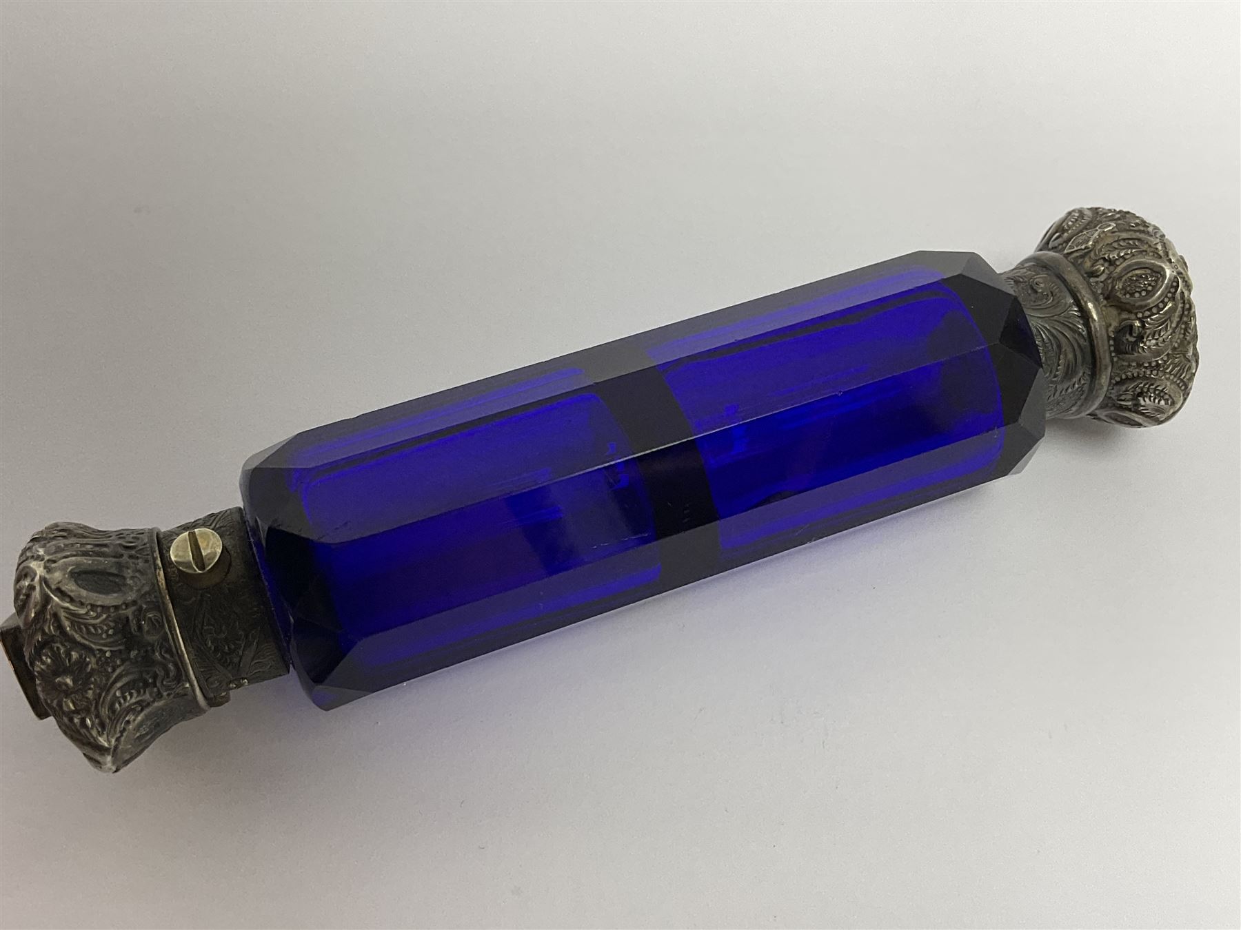 Late Victorian silver mounted ruby glass cylindrical scent bottle, with interior glass stopper, Birmingham 1900, makers mark worn, H8cm, together with a Victorian double ended cut blue glass perfume bottle mounted with white metal screw and flip caps decorated with foliate repousse detailing, H13.5cm