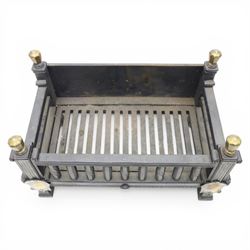 Arts and Crafts cast iron fire grate, rectangular backplate with plain upper rail over a slatted front of cylindrical tapering bars, flanked by square section uprights with fluted detail and scroll terminals, raised on stepped feet with shaped brass roundel mounts and brass finials to the corner posts