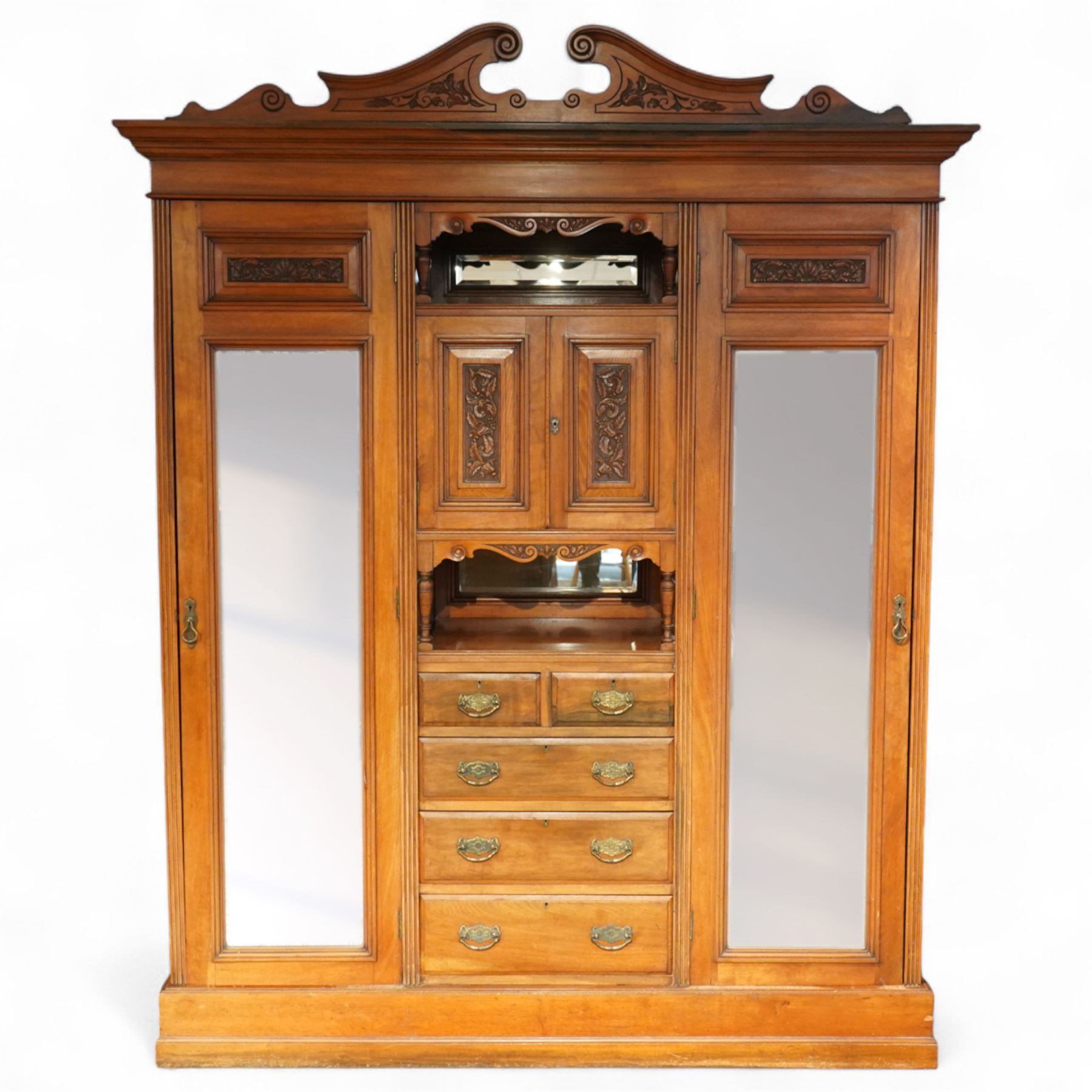 Edwardian walnut triple combination wardrobe, projecting pediment with pierced foliate scroll cresting above a central cupboard flanked by bevelled mirrored doors, the central section with twin panelled cupboard doors carved with foliate scrolls, over open recess with mirrored back and turned supports, above two short and three long drawers with brass handles, interior fitted with hanging rails and shelf, raised on plinth base