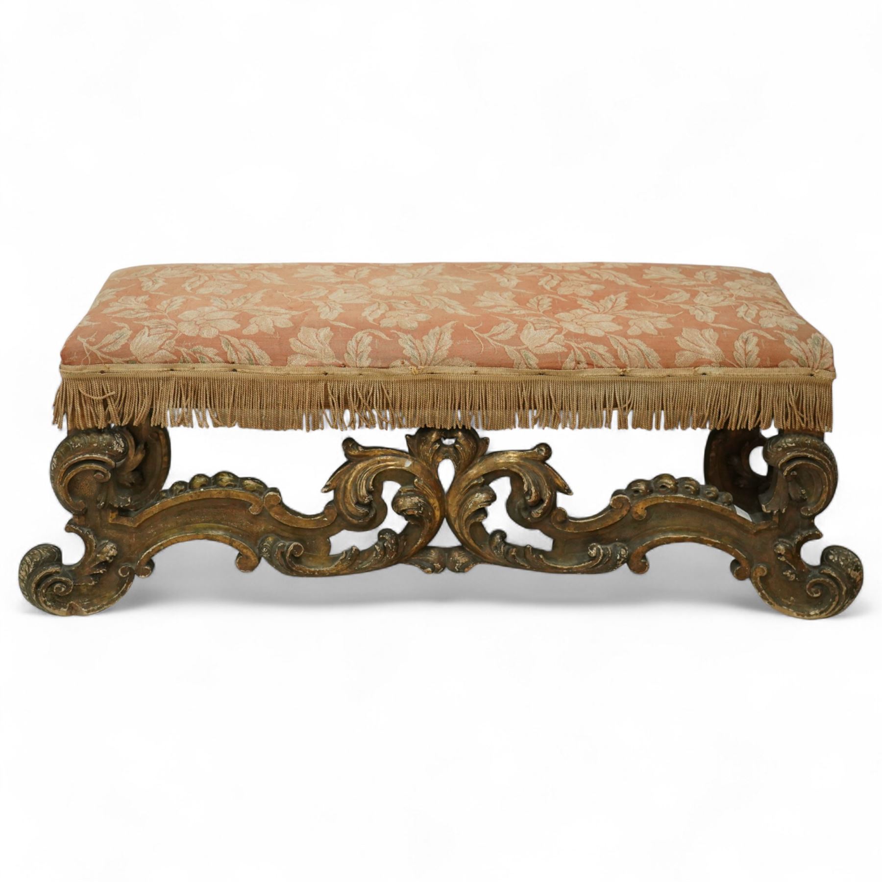 19th-century Rococo-style giltwood window seat, upholstered in floral-patterned coral-ground fabric with fringed border, the intricate scrolled supports united by pierced scrolling foliate stretchers with acanthus and C-scroll decoration