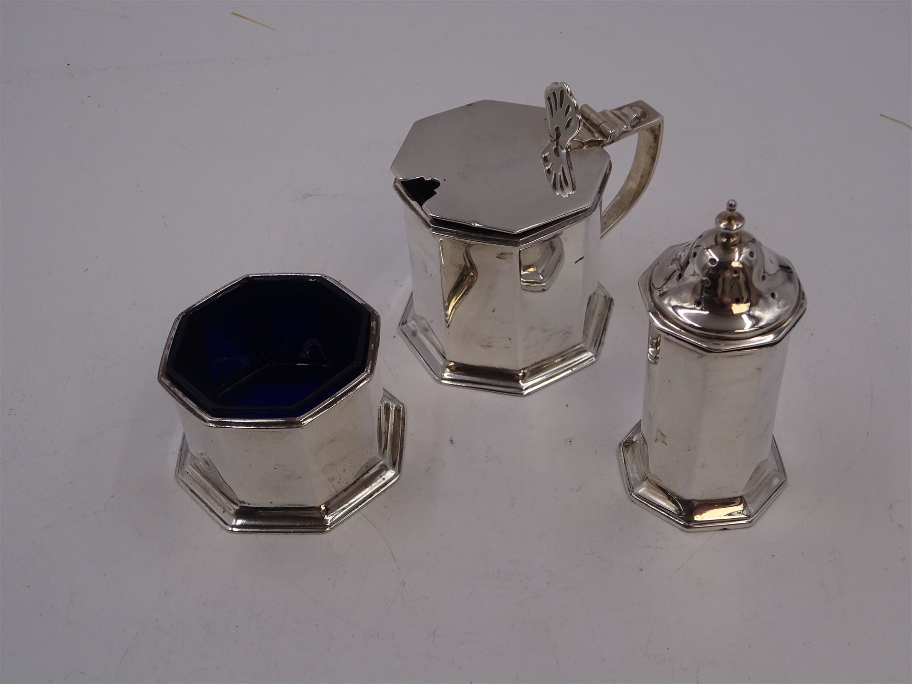 1920s silver three piece cruet set, comprising mustard pot and cover, pepper and open salt, each of faceted octagonal form, the mustard pot with pierced thumbpiece and capped handle, hallmarked Stokes & Ireland Ltd, Chester 1923 & 1924, max height 8.5cm, mustard and salt with original blue glass liners