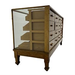 Early 20th century oak framed haberdashery shop counter, glass panel exterior enclosing sixteen graduating drawers with oak fronts and handles, raised on shaped tapering supports 