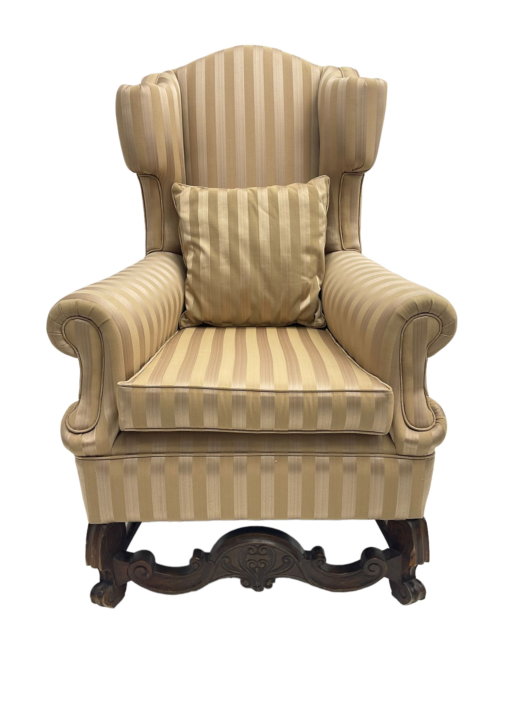 William and Mary style wingback armchair, upholstered in gold striped fabric, on walnut scroll carved supports joined by scroll and foliate carved middle rail, turned H-shaped stretchers, on castors