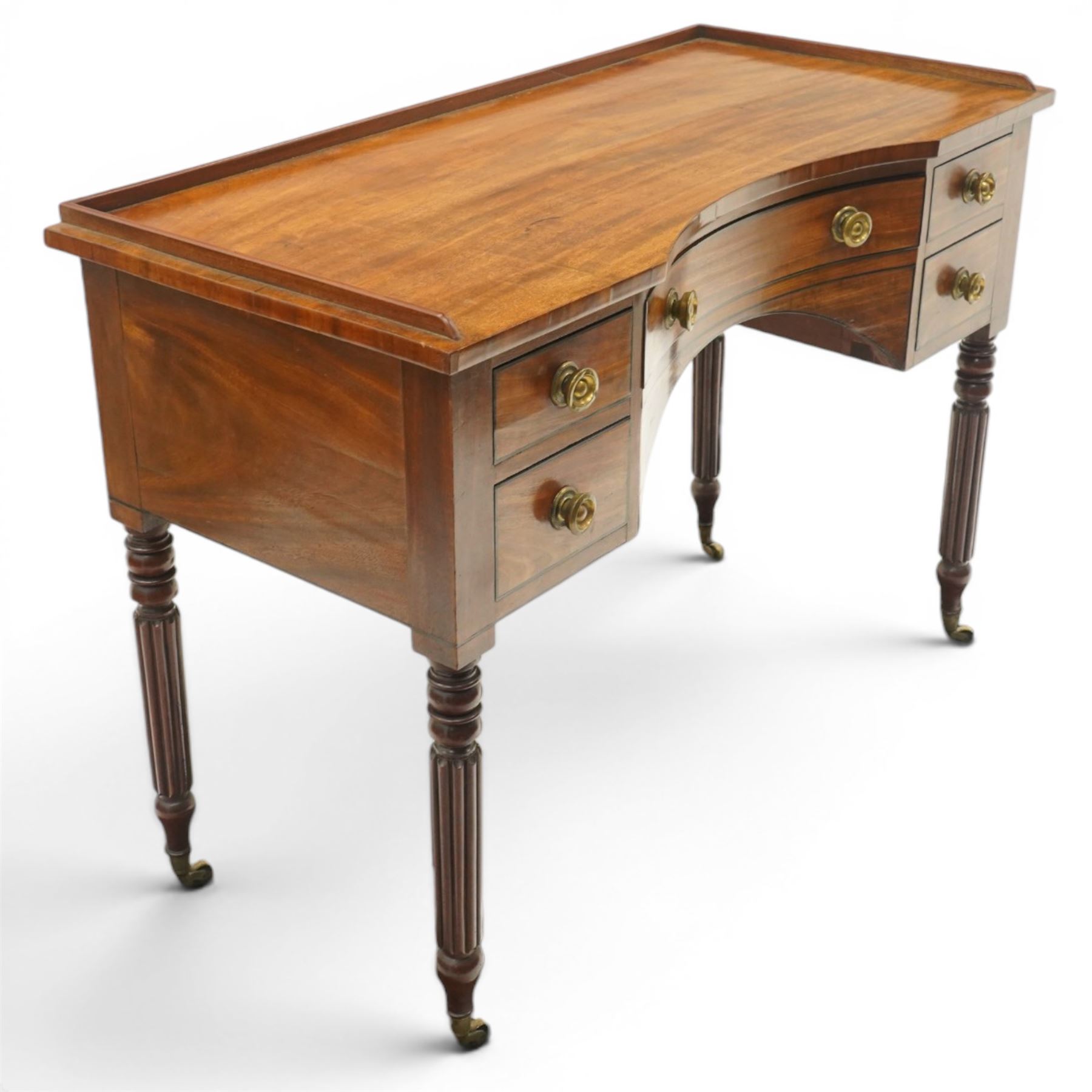 In the manner of Gillows - Regency period mahogany kneehole dressing table or writing desk, the shaped rectangular top with raised three-quarter gallery, fitted with central concave frieze drawer flaked by two graduating drawers to each side, each with ebony stringing, raised on turned and reeded supports terminating to brass cups and castors