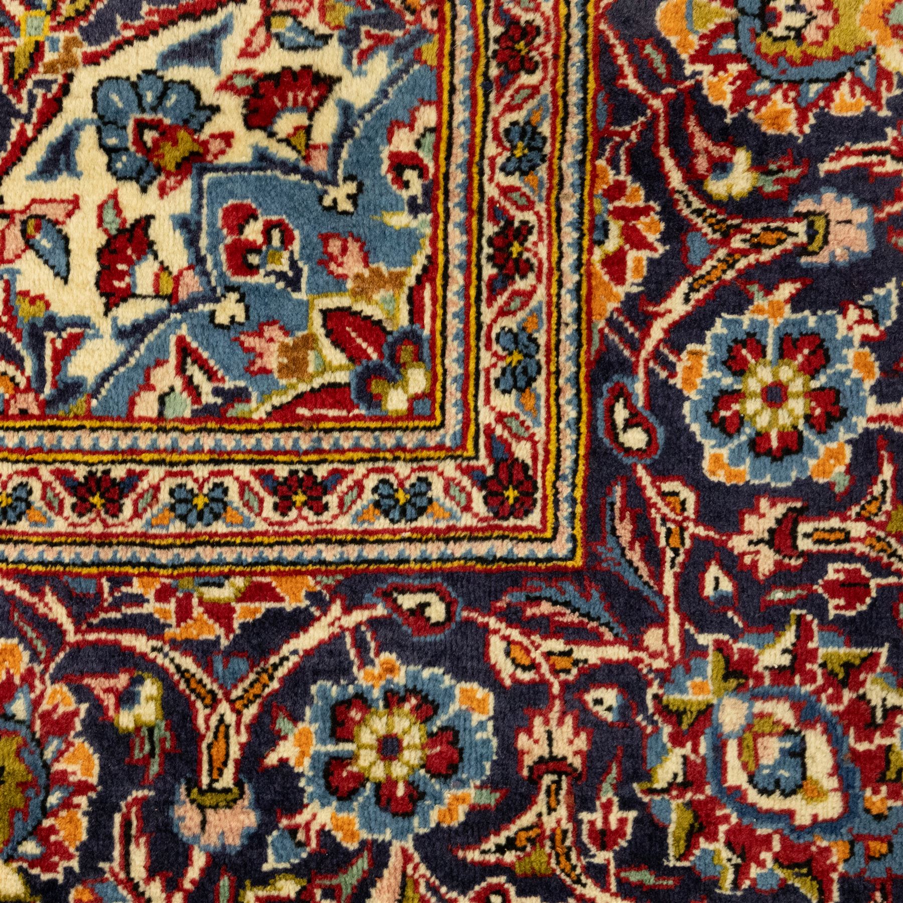 Central Persian Kashan hand-knotted carpet, crimson red central field with central floral medallion with floral vine-work and palmettes, within dense floral spandrels and a wide navy blue border decorated with continuous foliate motifs and multiple guard borders, with cotton fringe ends  