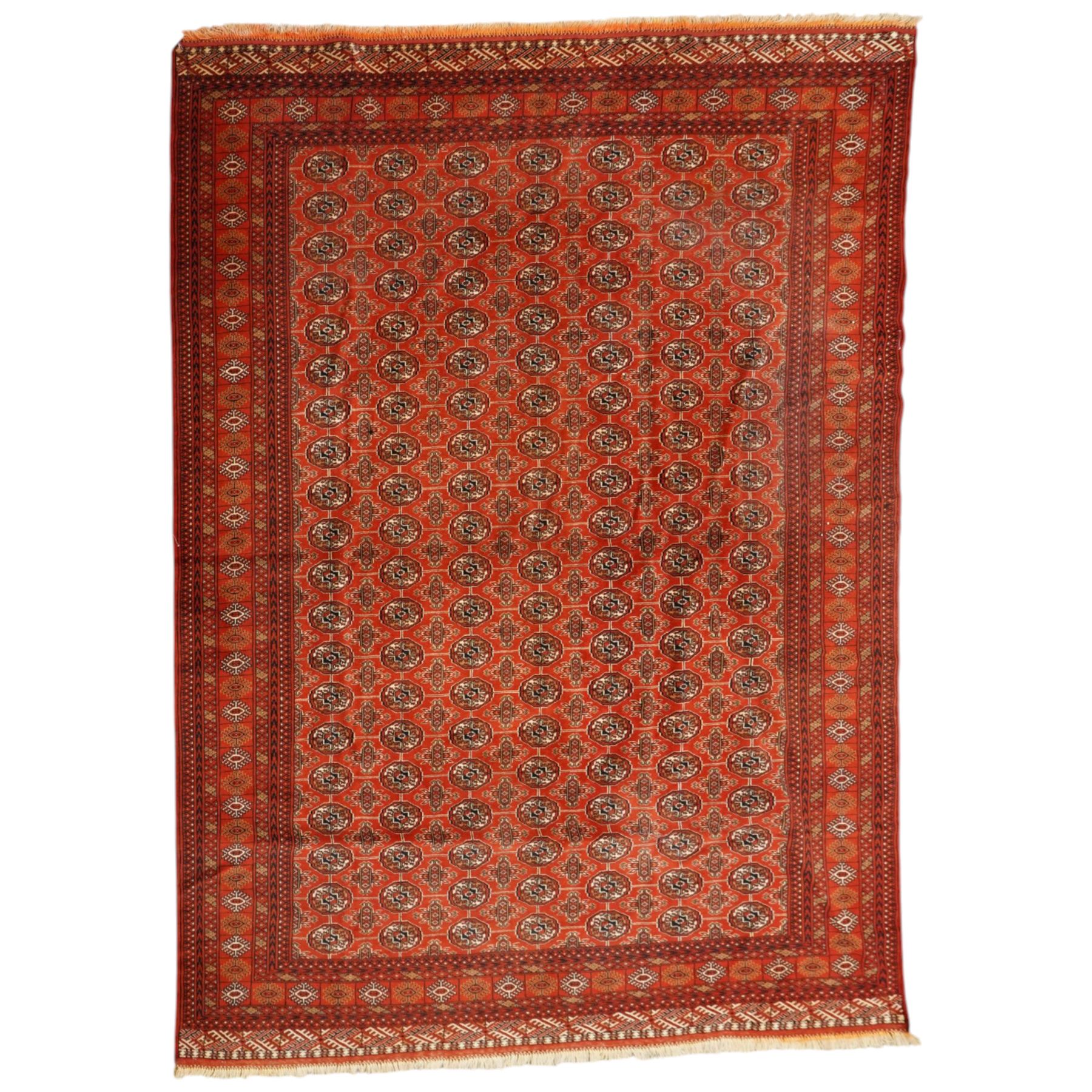Turkmen hand-knotted carpet, madder red field, with an all-over design of row of Tekke guls, each enclosing stylized cruciform and geometric devices in tones of ivory, dark brown, and indigo, principal border of stylized geometric motifs, flanked by guard border with angular latch hook geometric designs, with cotton fringe ends 
