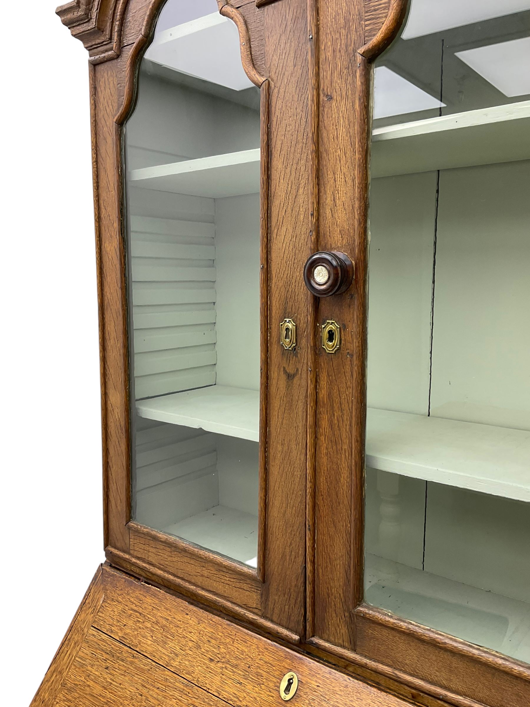 George III oak bookcase on bureau, the double arched top enclosed by two shaped glazed doors, fall-front concealing fitted interior, over four graduating cock-beaded drawers, on bracket feet