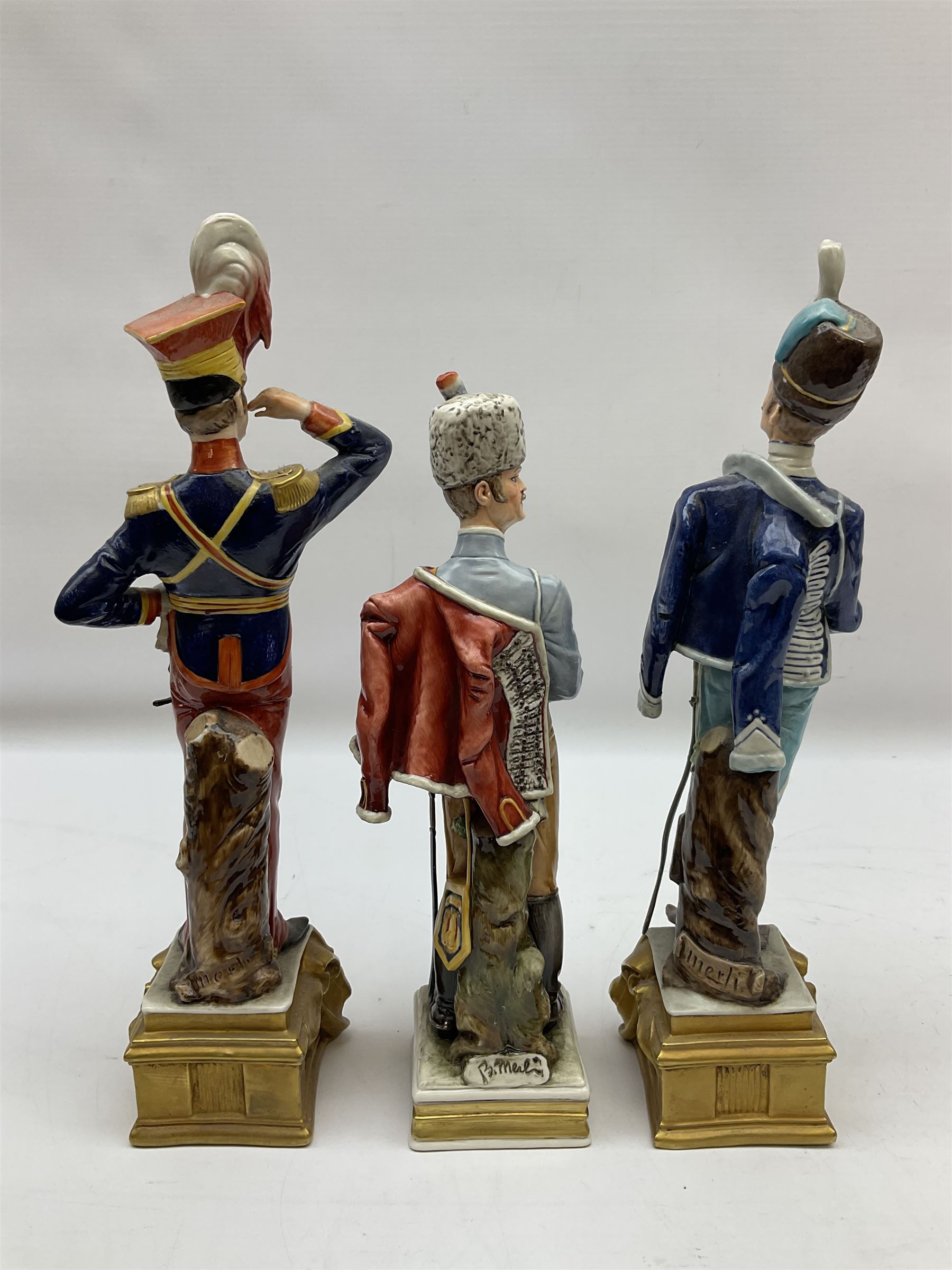 Group of seven Capodimonte figures of soldiers, to include four Bruno-Merli examples including Officer of the 12th Lancers 1820 and Captain of the 18th Hussars 1815, both raised upon ornate gilt plinths bearing British monarch motto in Latin 'Dieu et mon droit', and three further similar, all with crowned N mark beneath, tallest H32cm