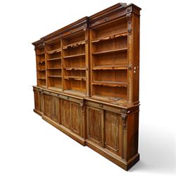 Large early 19th century Country House mahogany breakfront library bookcase, the projecting moulded cornice over a frieze applied with carved flowerhead paterae, the open panelled upper section fitted with adjustable shelves and shaped valances, divided by moulded pilasters with acanthus-carved corbels, the projecting base enclosed by eight panelled cupboard doors with central turned canted pilaster, on plinth base