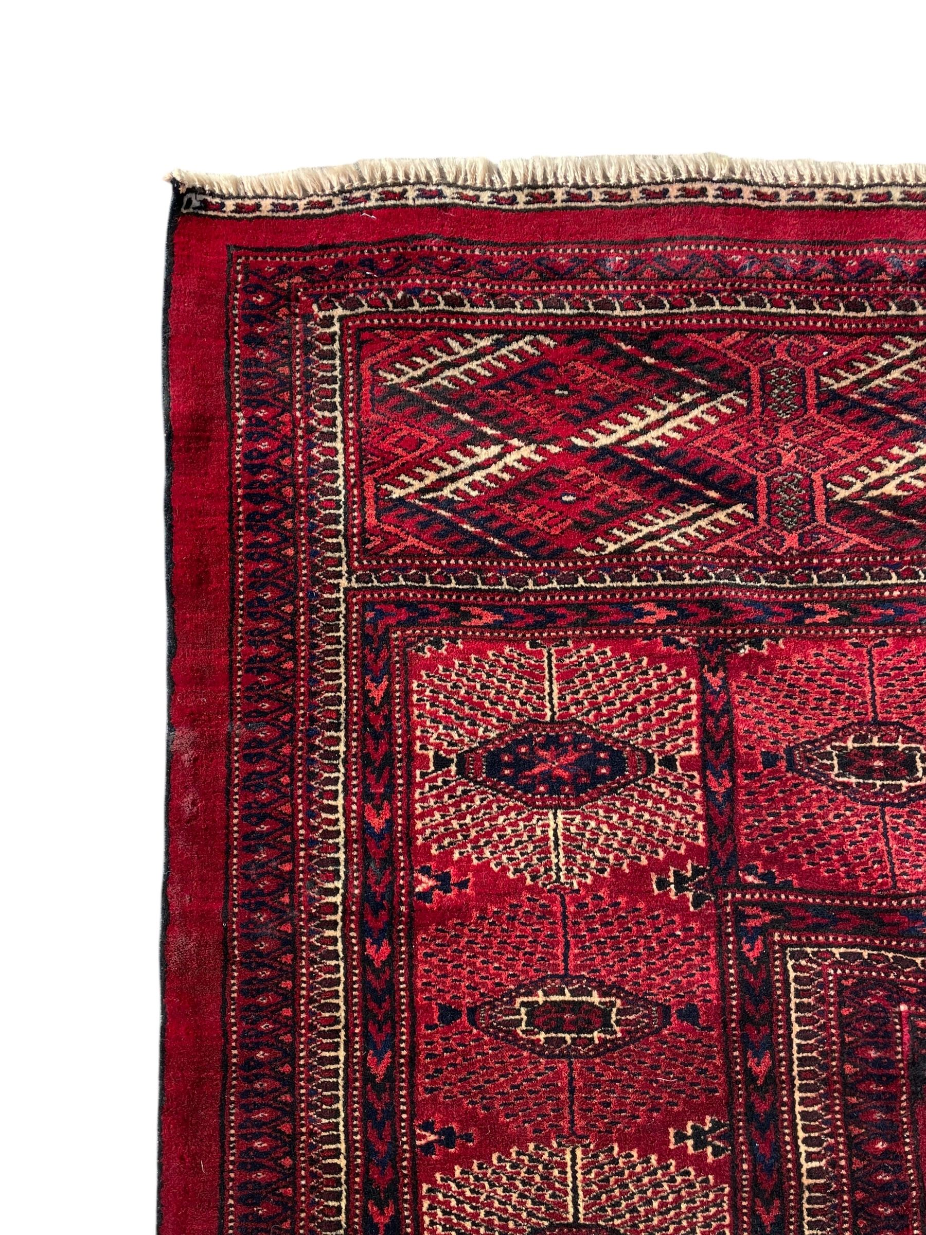 Afghan Bokhara red ground carpet, the field decorated with four rows of Gul motifs interspersed with Hac motifs, the border with lattice pattern panels within small geometric guard stripes, geometric patterned end panels