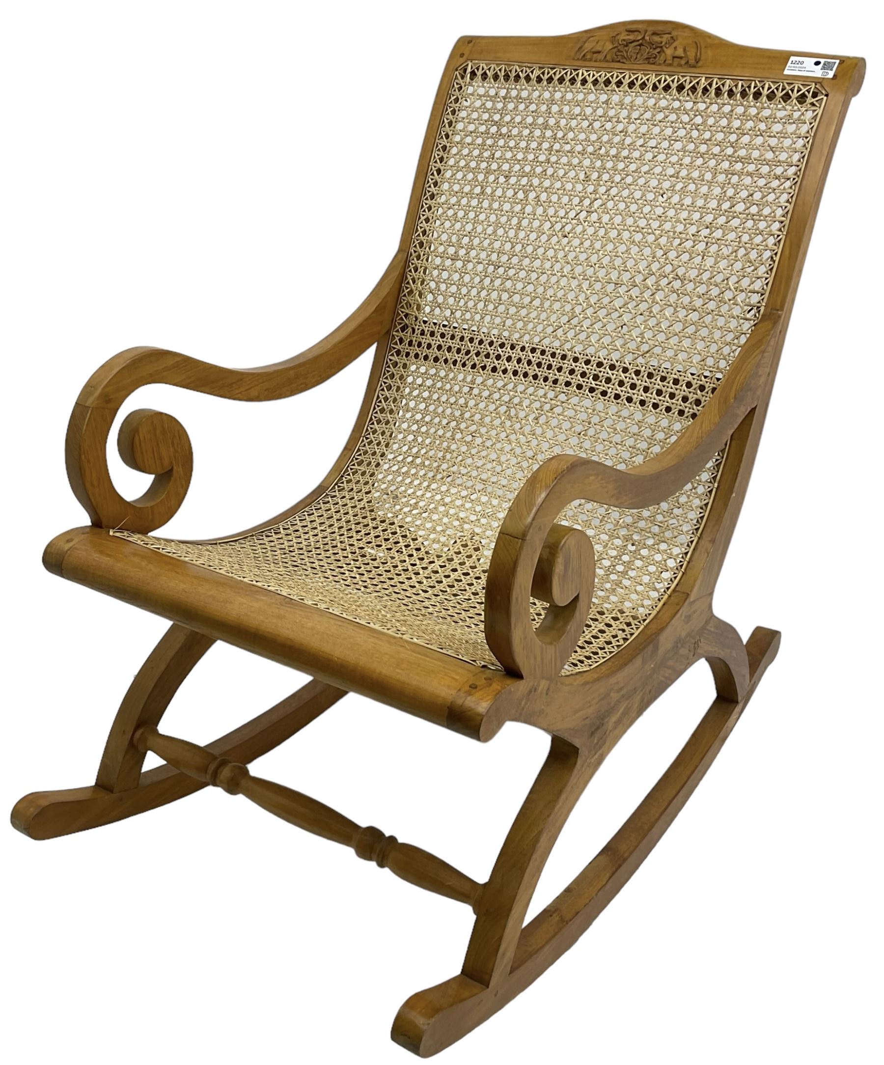 Late 20th century teak framed plantation style rocking chair, cresting rail carved with two elephants with upraised trunks, cane back and seat with scrolled arm terminals, rocker base united by turned stretcher