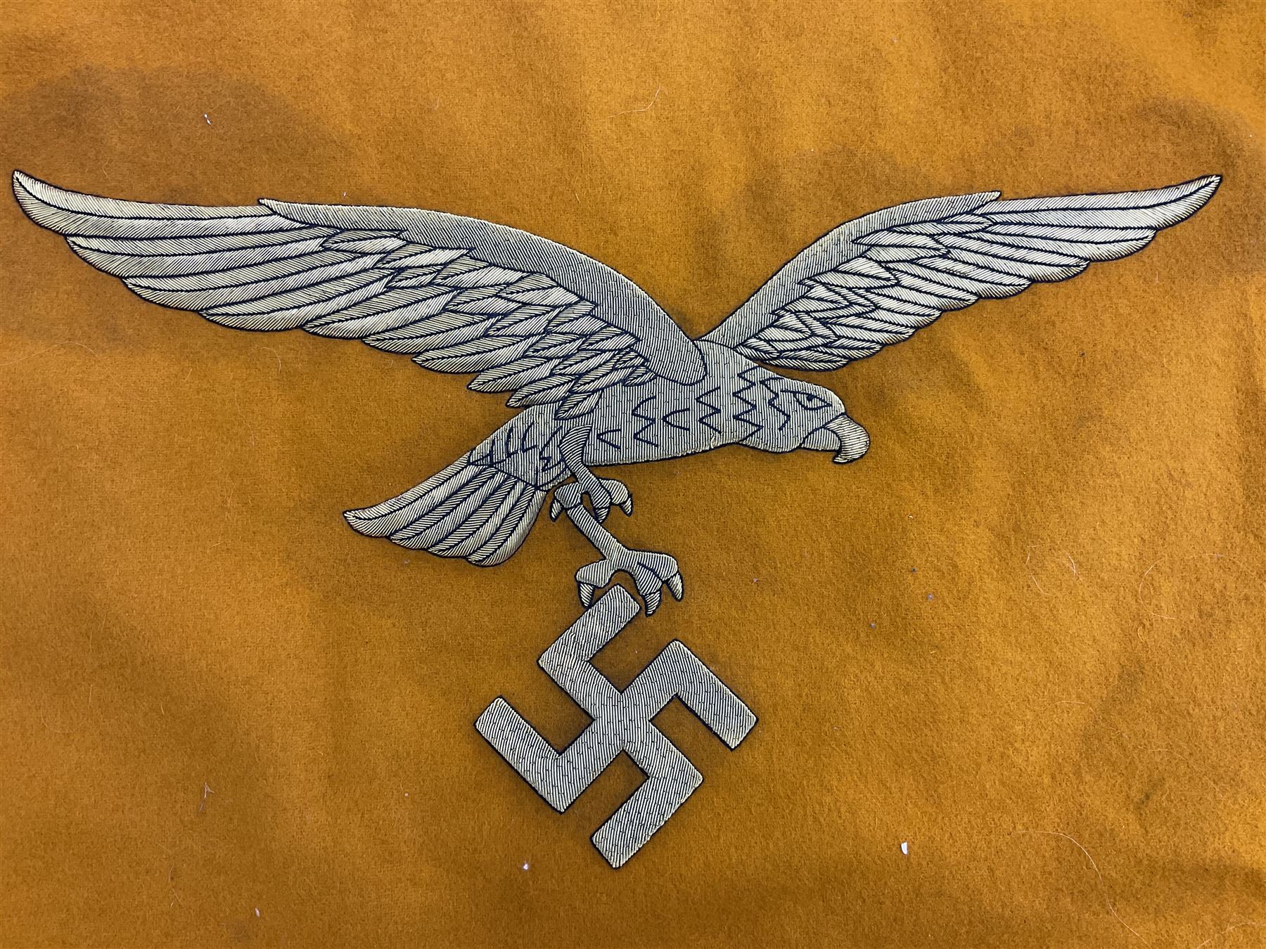 Two replica German Luftwaffe trumpet banners, one heavily silver wire embroidered with Luftwaffe eagle in a wreath, on an amber, white and black rayed background, , with silver wire tasselled border and four leather hanging loops, approx. 50 x 49cm; the other slightly larger heavily gold wire embroidered with the Luftwaffe eagle on a plain amber ground with gold wire tasselled border and two hanging loops (2)