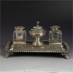 George IV silver desk stand, of rectangular form with rounded corners and oblique gadrooned rim, upon four embossed pad feet, with central pedestal dish of part fluted form with domed lid engraved with crest and with bud finial, with two recesses either side housing faceted glass inkwells, each with silver collars, hallmarked John & Thomas Settle, Sheffield 1825 & 1827, H11cm