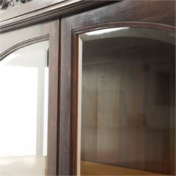 Large and impressive 19th century heavily carved mahogany display cabinet, the ornate pediment carved with a central scalloped and foliate rocaille motif with extending acanthus leaves and egg-and-dart moulding flanked by acanthi finials, over a central cartouche with carved bow-and-arrow motif and trailing scrollwork, the bookcase enclosed by two glazed doors with bevelled glass sides, the base with a gadrooned upper edge over a central rocaille ornament with scrolling acanthus leaves, the panelled frieze with further foliate motifs, raised on large carved snarling lion mask supports with floral garlands and claw feet, united by a concave rectangular platform base with gadrooning egg-and-dart moulding