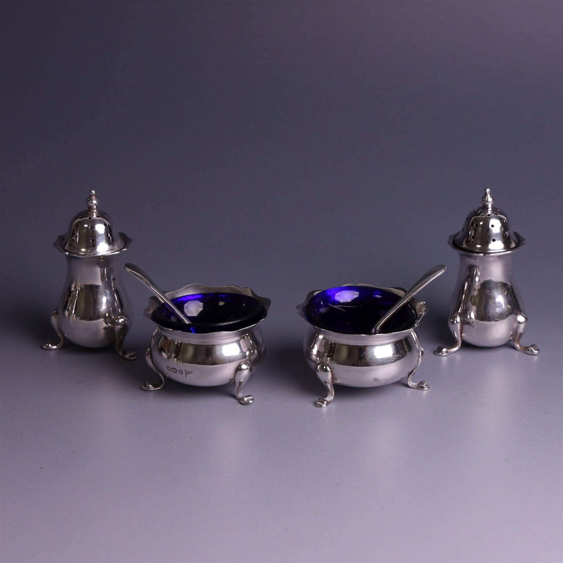 Early 20th Century four piece silver cruet set, comprising a pair of pepper shakers and a pair of open salts, each of circular form with shaped rims and upon three pad feet, hallmarked Walker & Hall, Sheffield 1912, the open salts with two silver plated salt spoons and blue glass liners 