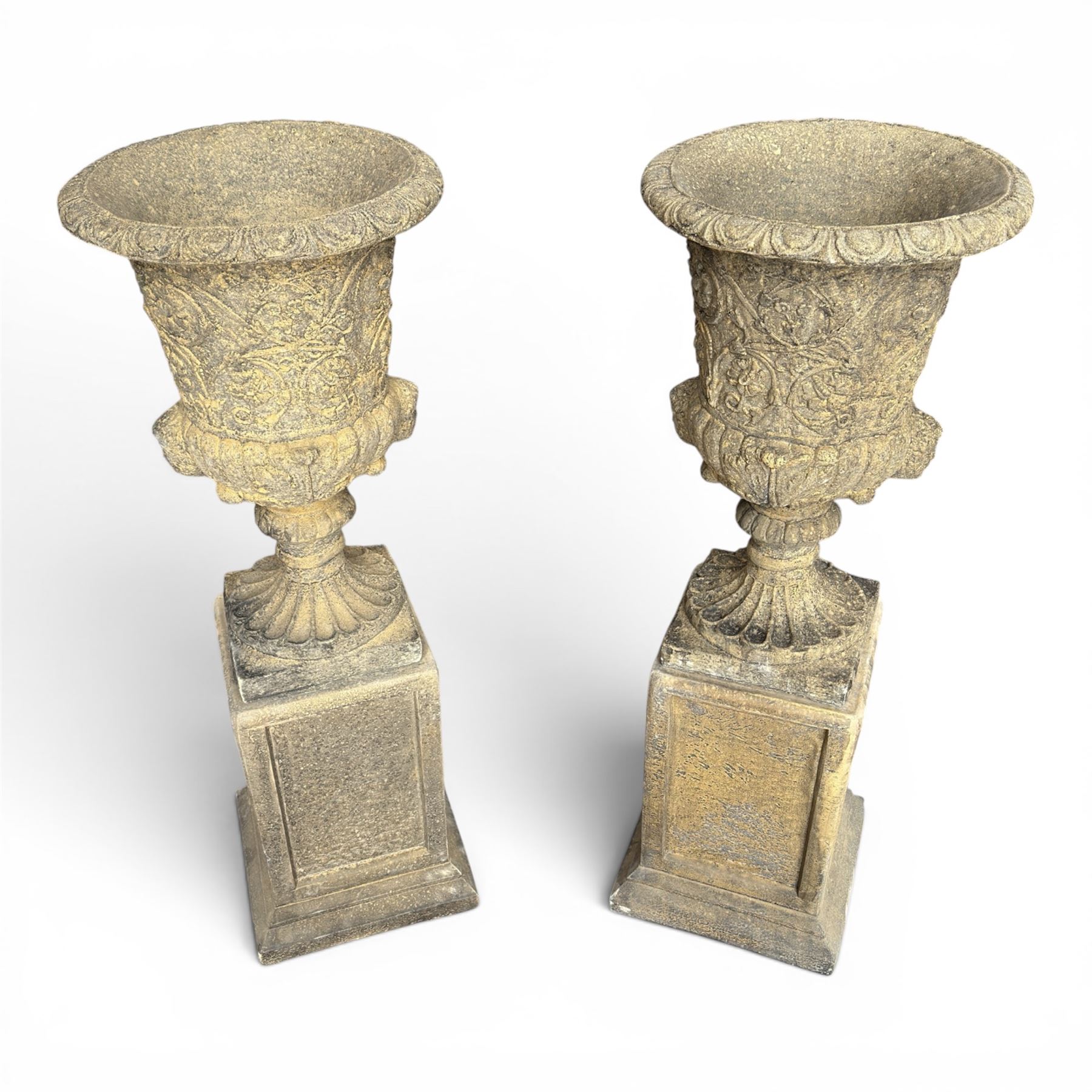Pair of George III design antiqued composite stone garden urns of campana form, the egg-and-dart everted rim over a waisted body relief moulded with scrolling fruiting foliage and flowerheads, the gadrooned underbelly with twin mask ornaments, the knopped stems on fluted socles, raised on faux panelled square pedestal bases